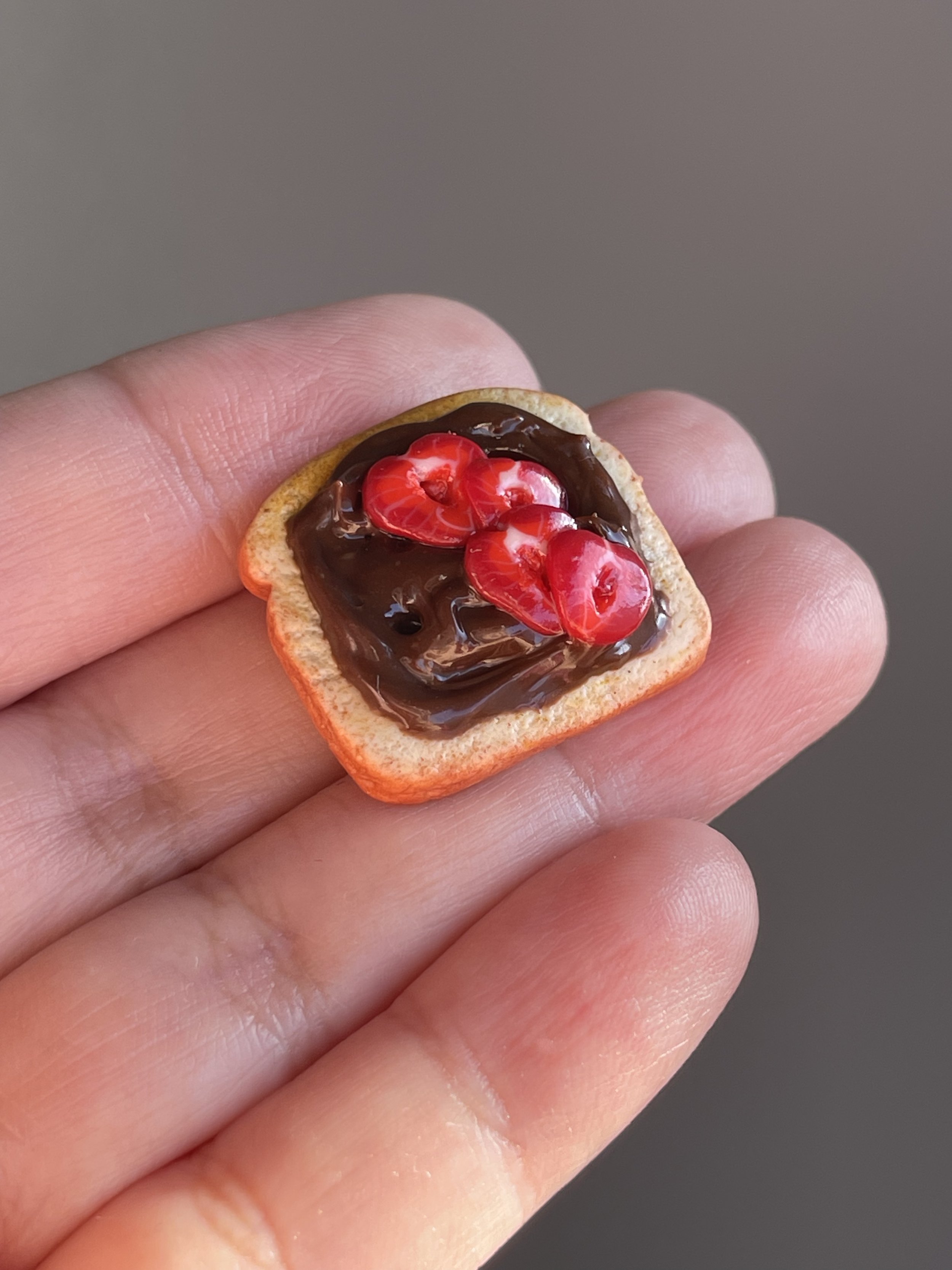 A hand holding a miniature polymer clay hazelnut spread and sliced strawberries toast magnet.