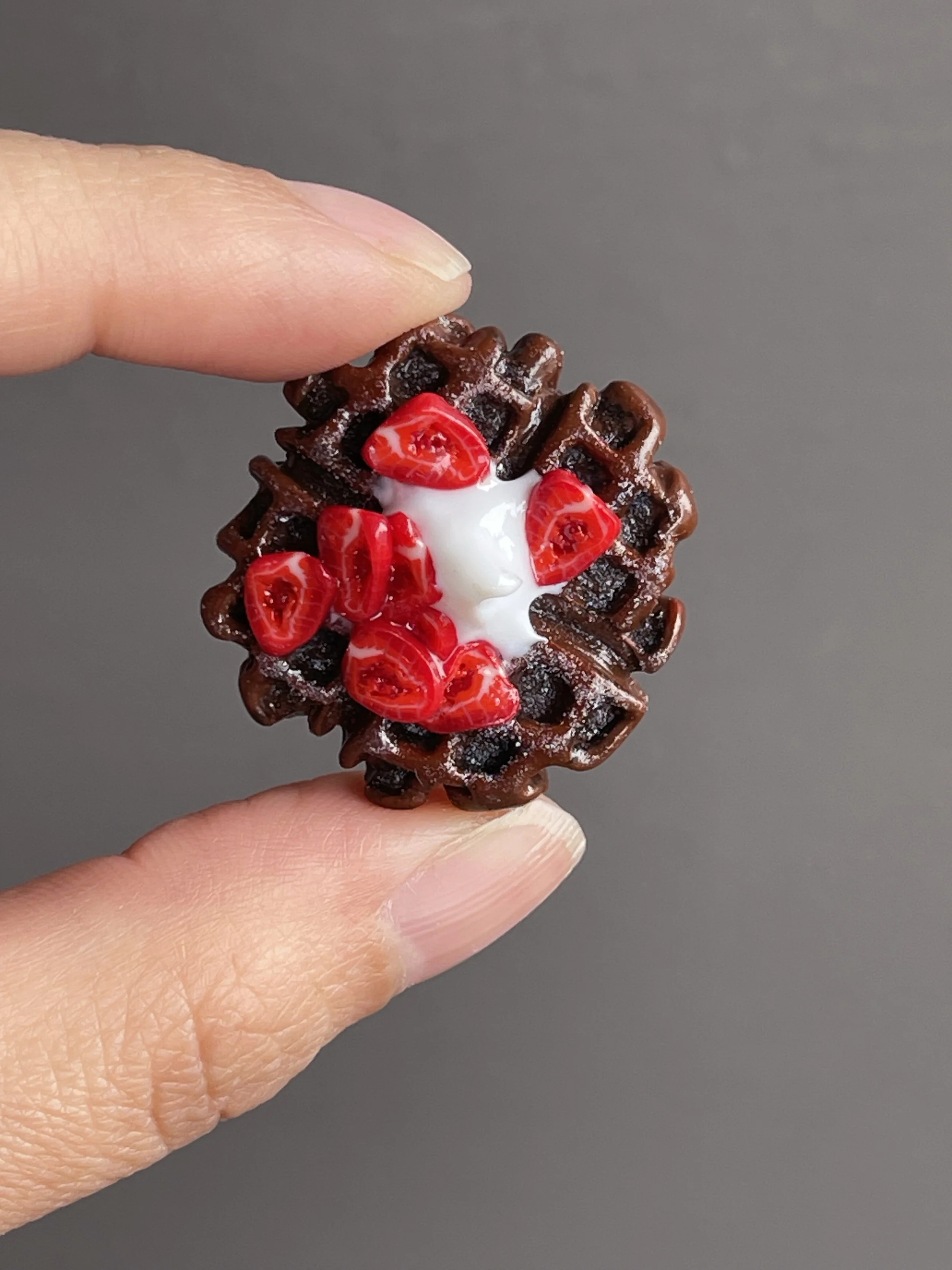 Fingers holding a miniature polymer clay chocolate waffle magnet topped with strawberries and whipped cream.