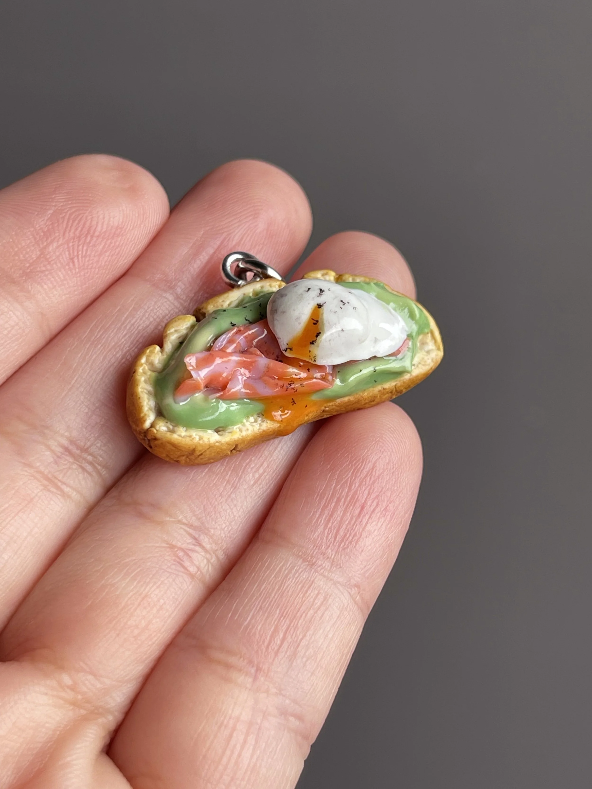 A hand holding a miniature polymer clay avocado toast keychain with smoked salmon, avocado mash, and a runny poached egg.