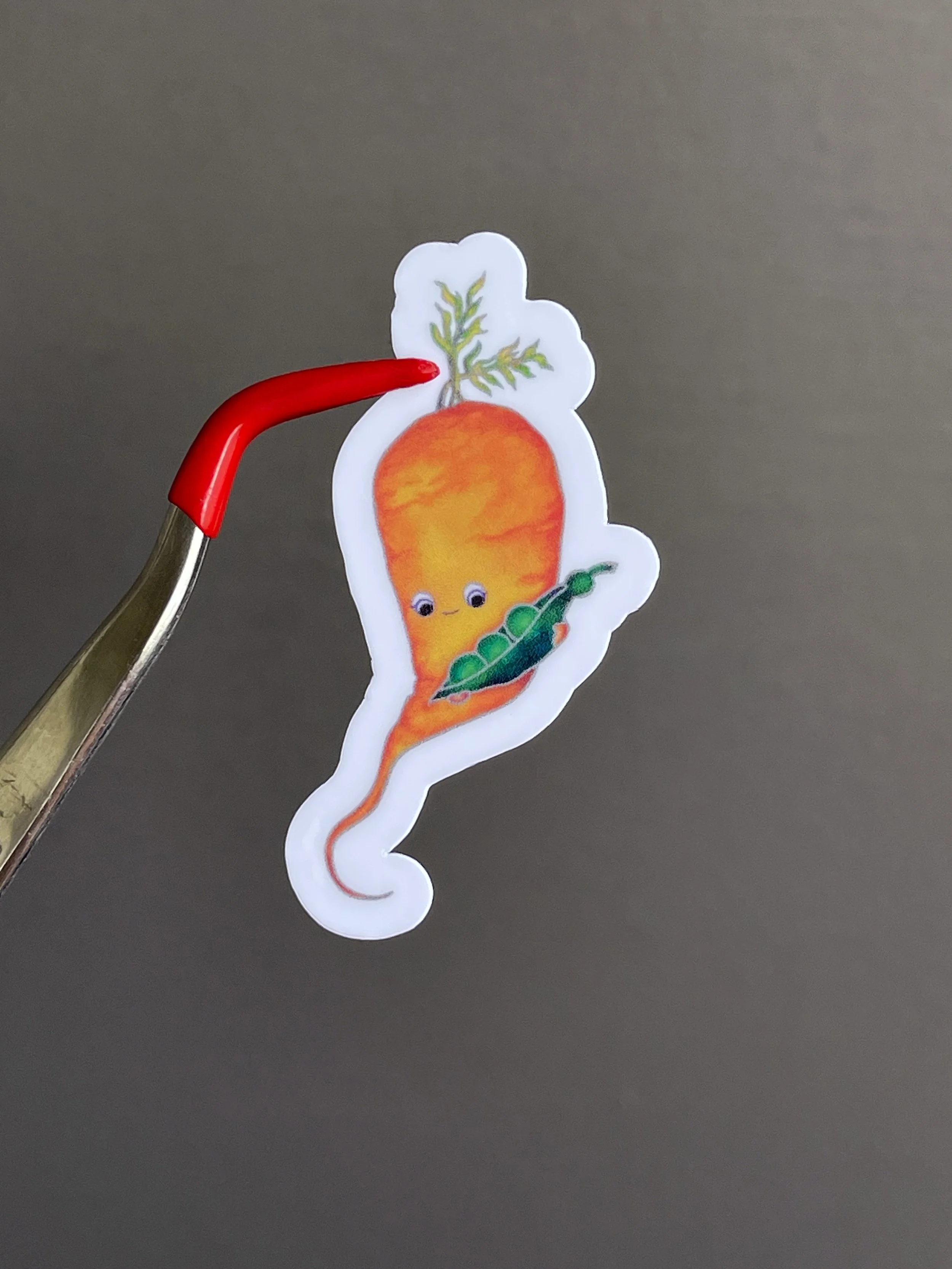 Red tip bent tweezer holding a sticker of a cute carrot character holding an open pea pod.