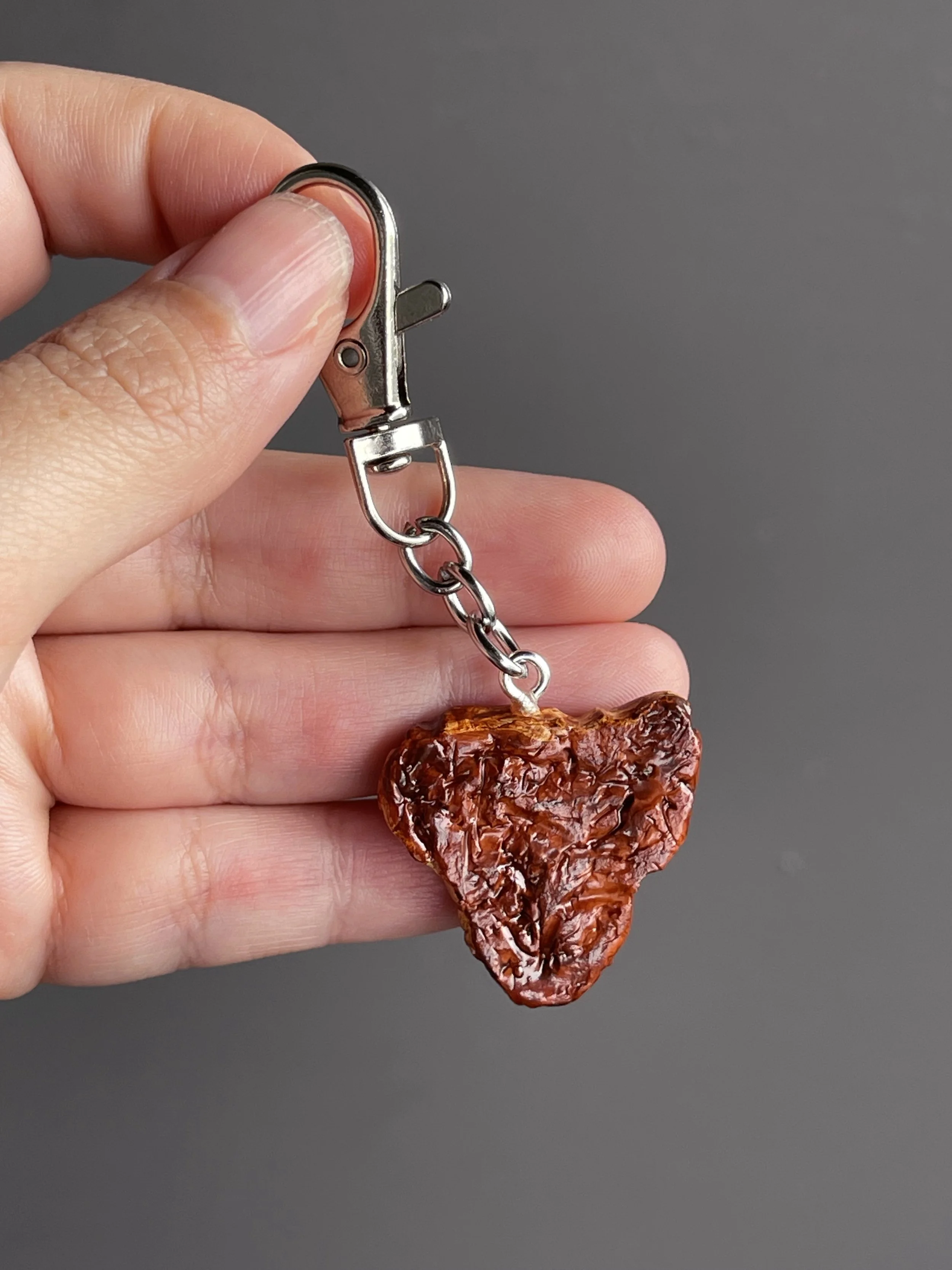 A hand holding a realistic miniature polymer clay t-bone steak keychain showing the back of the piece.