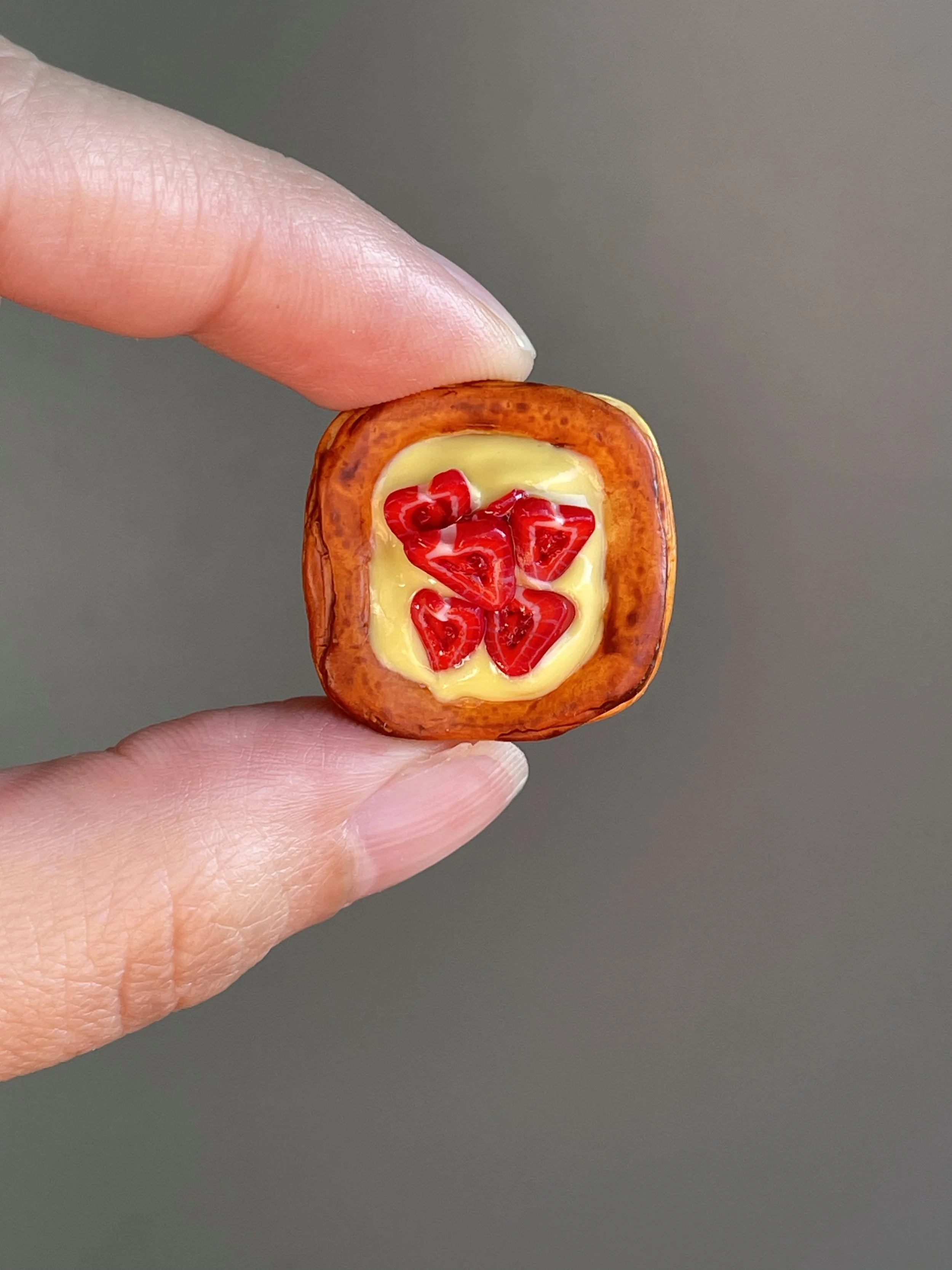 Fingers holding a miniature polymer clay danish with yellow custard and strawberry slices magnet.