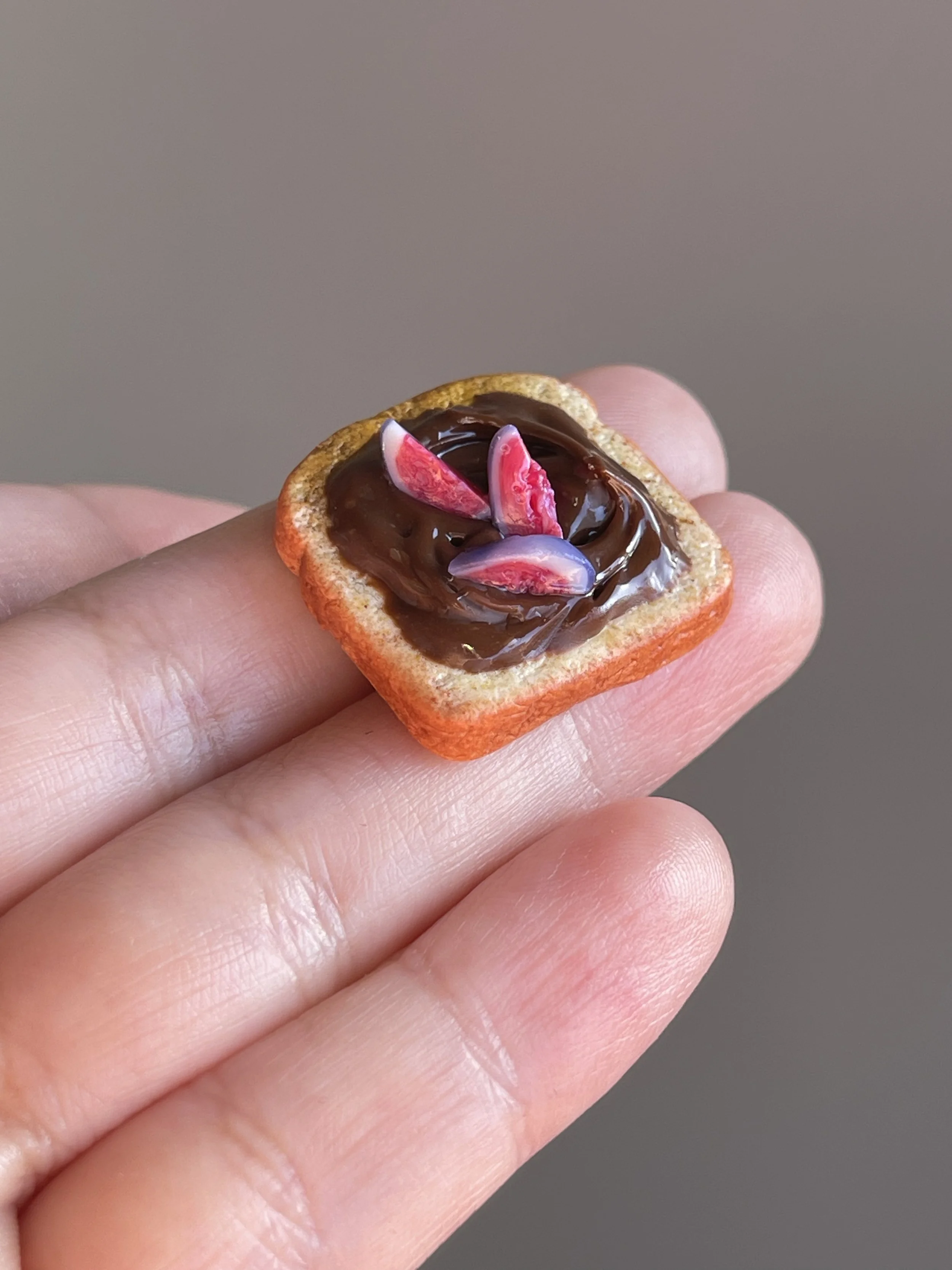 A hand holding a miniature polymer clay hazelnut spread and figs toast magnet.