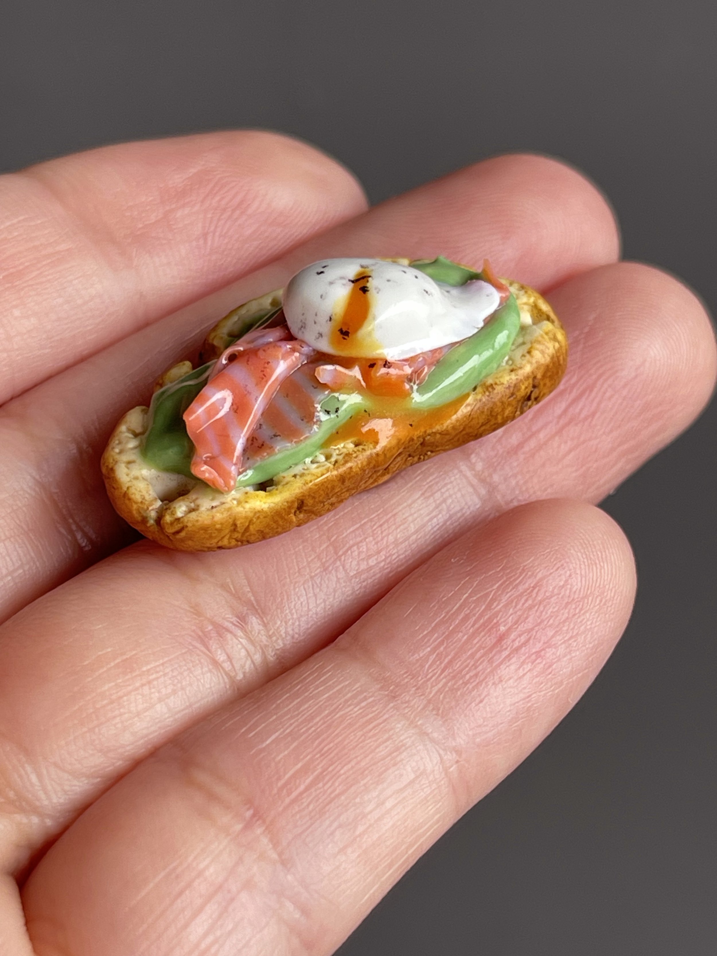A hand holding a miniature polymer clay avocado toast magnet with smoked salmon, avocado mash, and a runny poached egg.