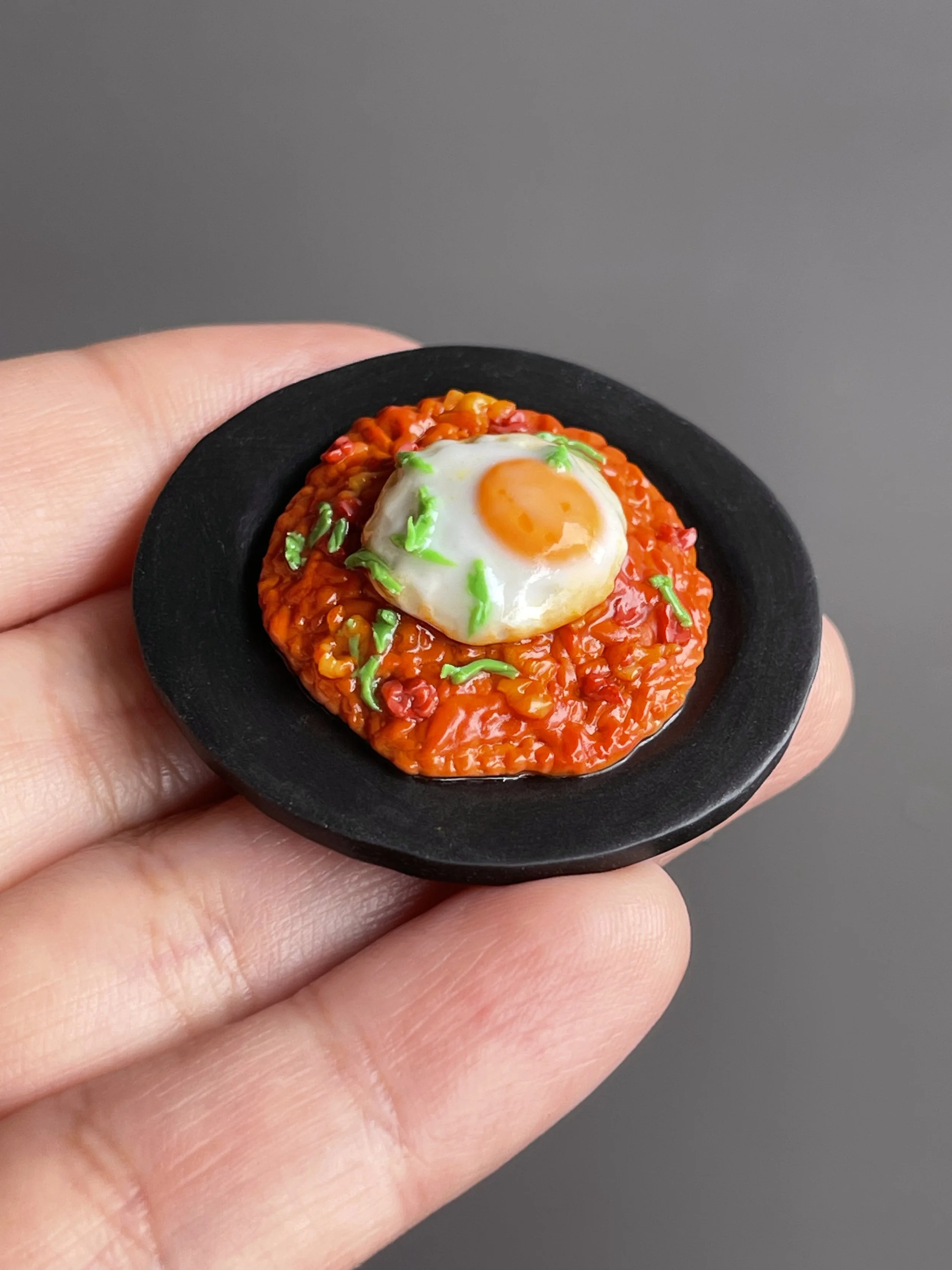 A hand holding a miniature polymer clay kimchi fried rice magnet topped with a fried egg and green onions atop a black plate.