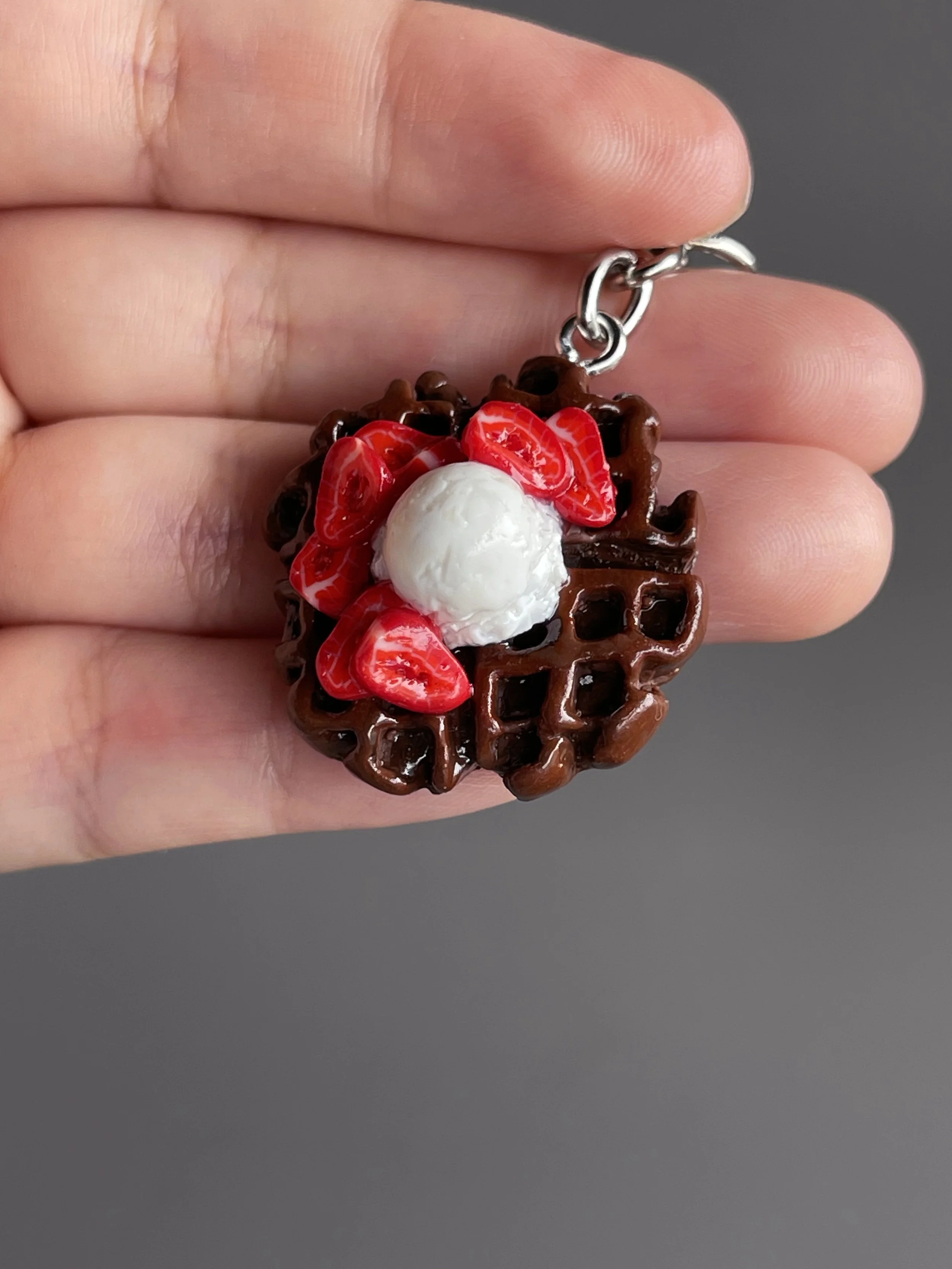 A hand holding a miniature polymer clay chocolate waffle keychain with strawberry slices & ice cream toppings.