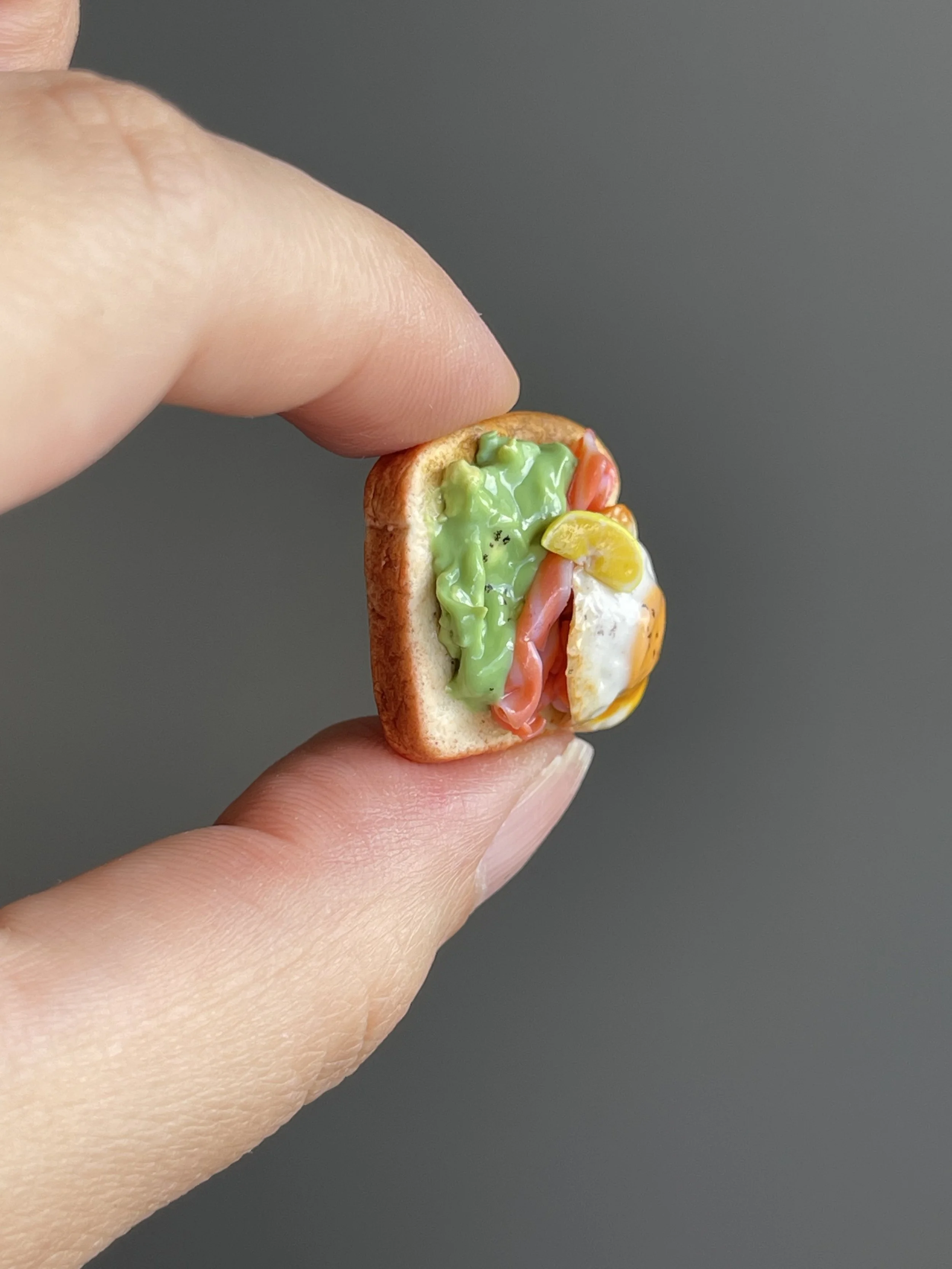 Fingers holding a miniature polymer clay egg, smoked salmon, avocado mash toast magnet with a lemon slice garnish.