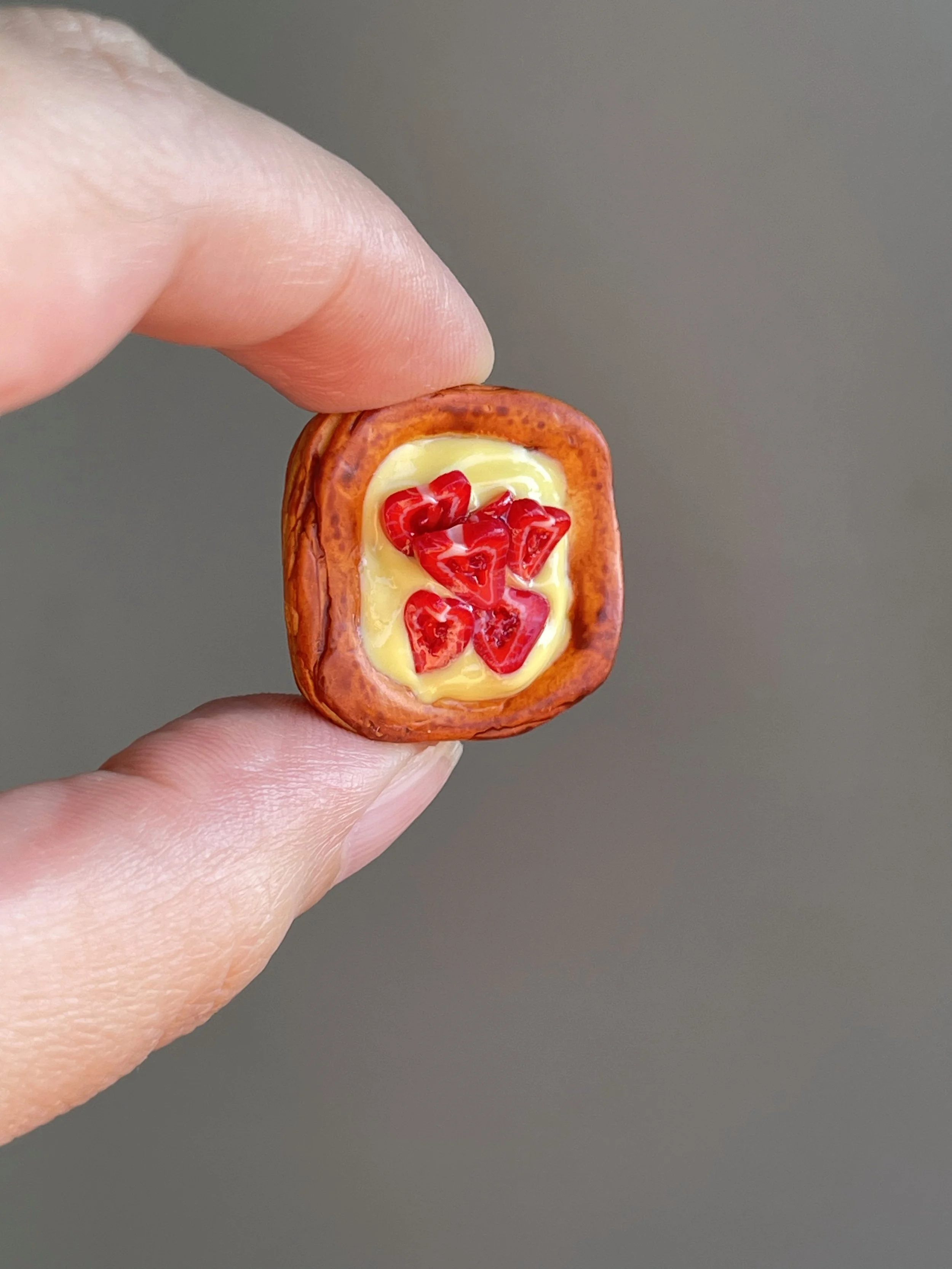 Fingers holding a miniature polymer clay danish with yellow custard and strawberry slices magnet.