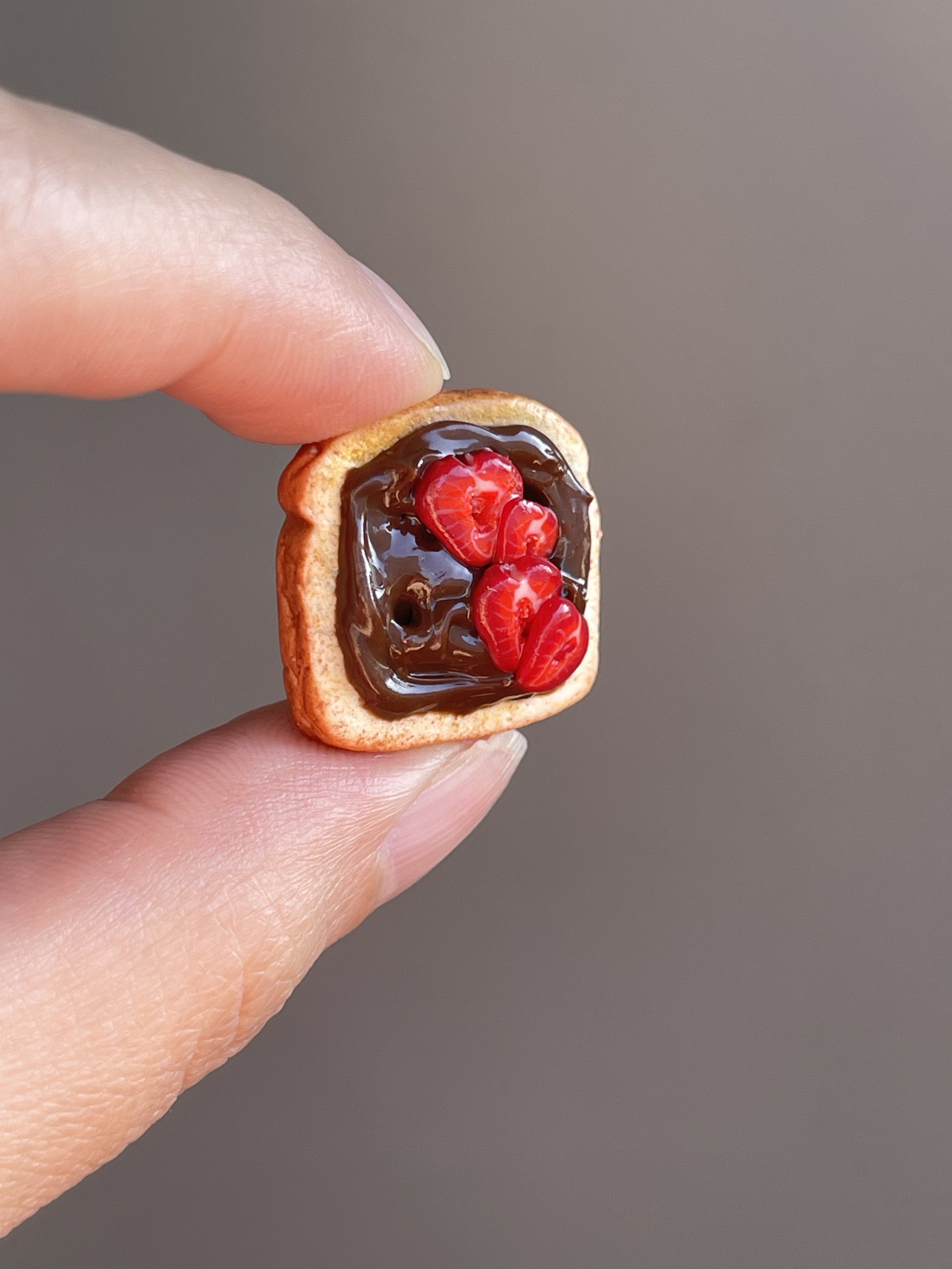 Fingers holding a miniature polymer clay hazelnut spread and sliced strawberries toast magnet.