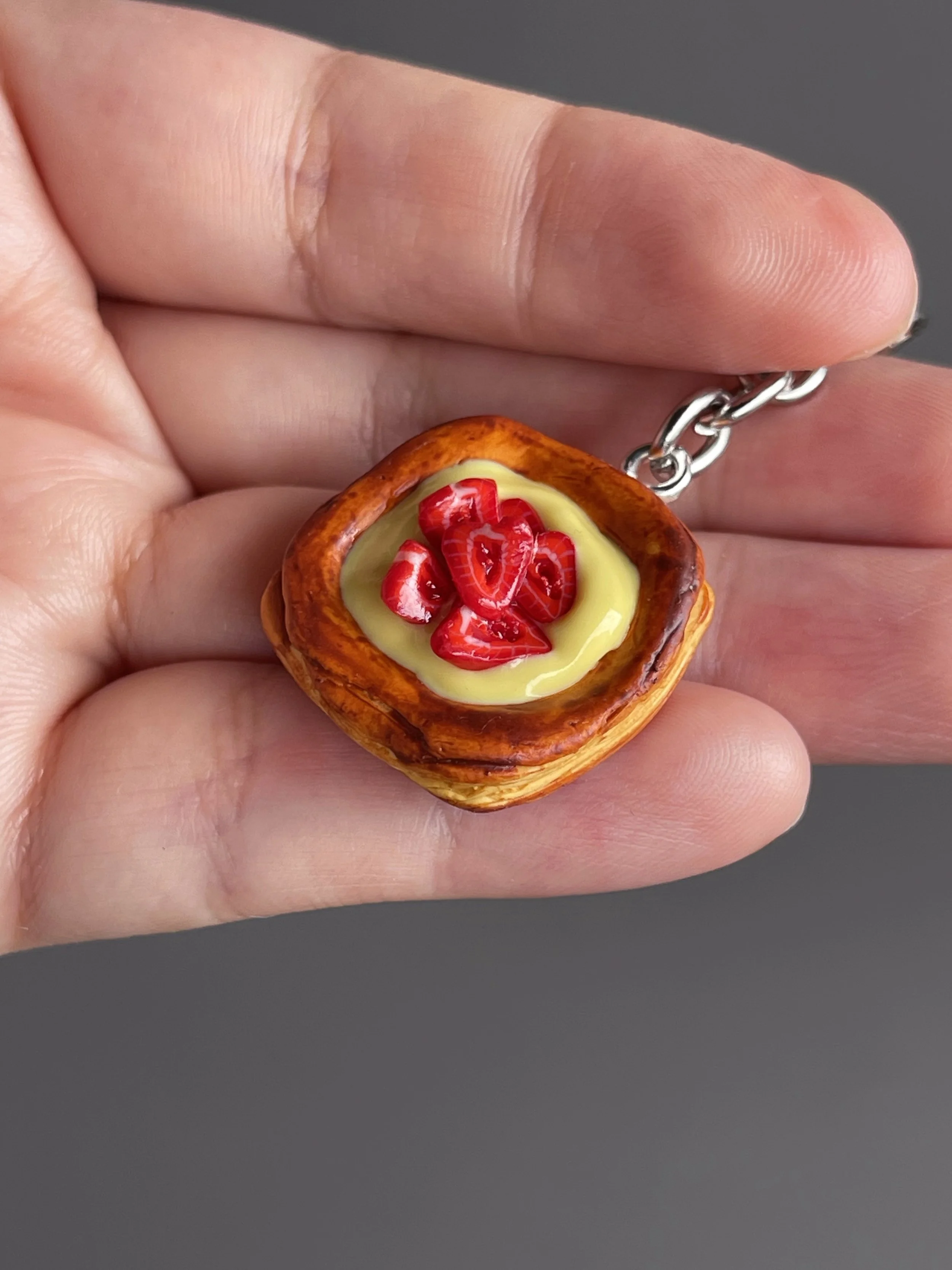 A hand holding a miniature danish pastry with yellow custard and strawberry slices keychain.