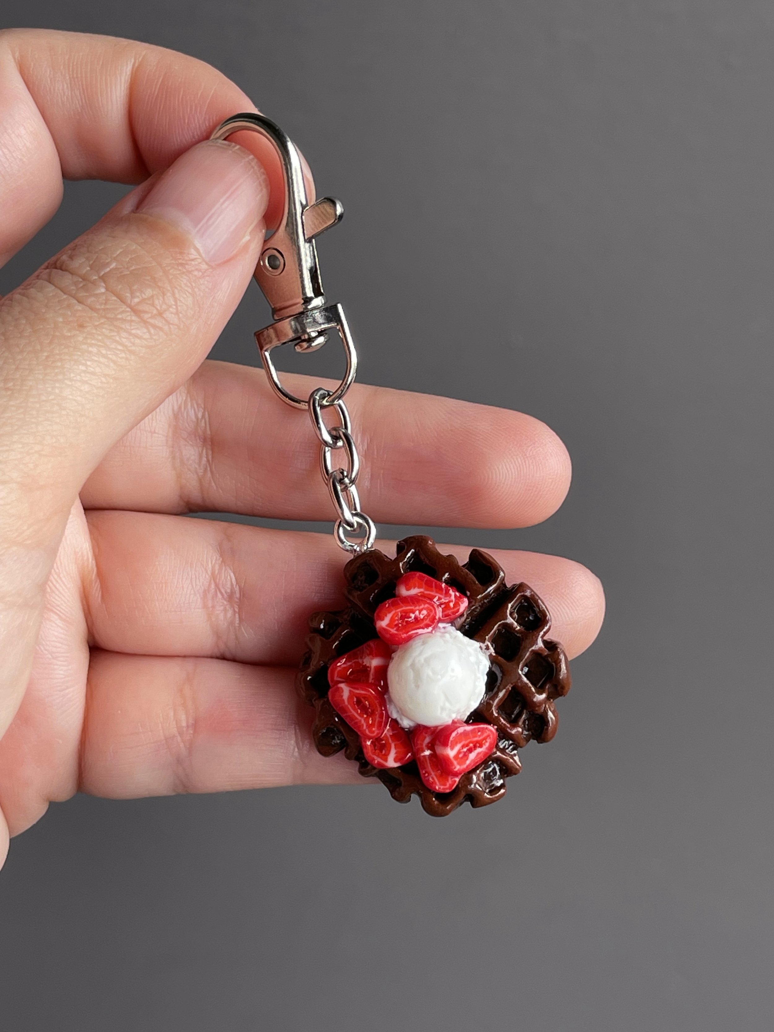 A hand holding a miniature polymer clay chocolate waffle keychain with strawberry slices & ice cream toppings.