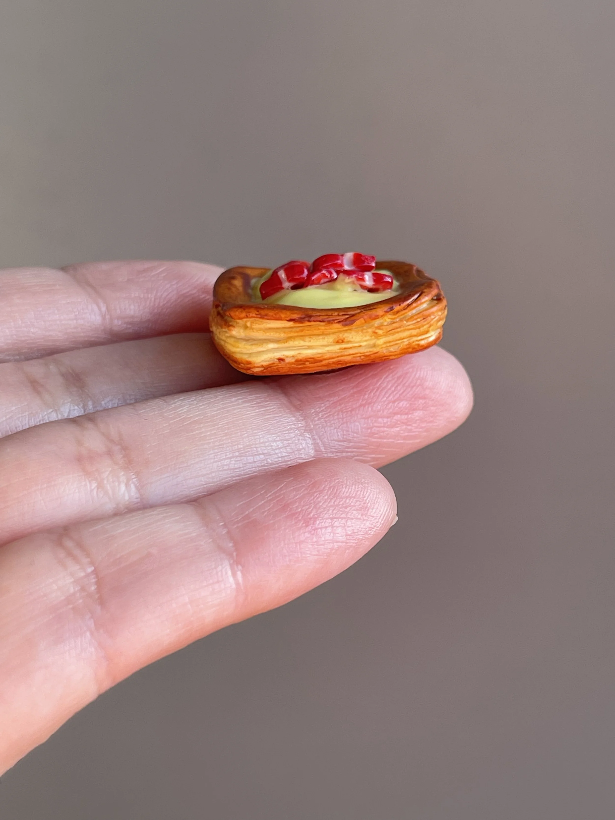A hand holding a miniature polymer clay danish with yellow custard and strawberry slices magnet.