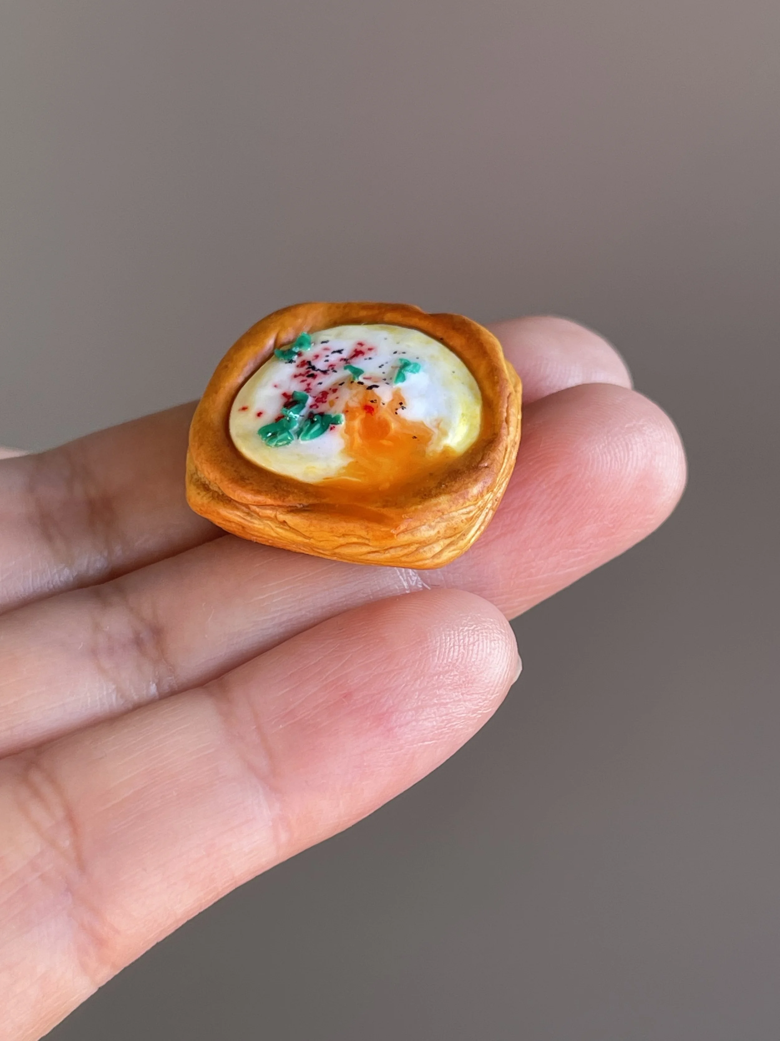 A hand holding a miniature polymer clay egg danish magnet with runny egg yolk.