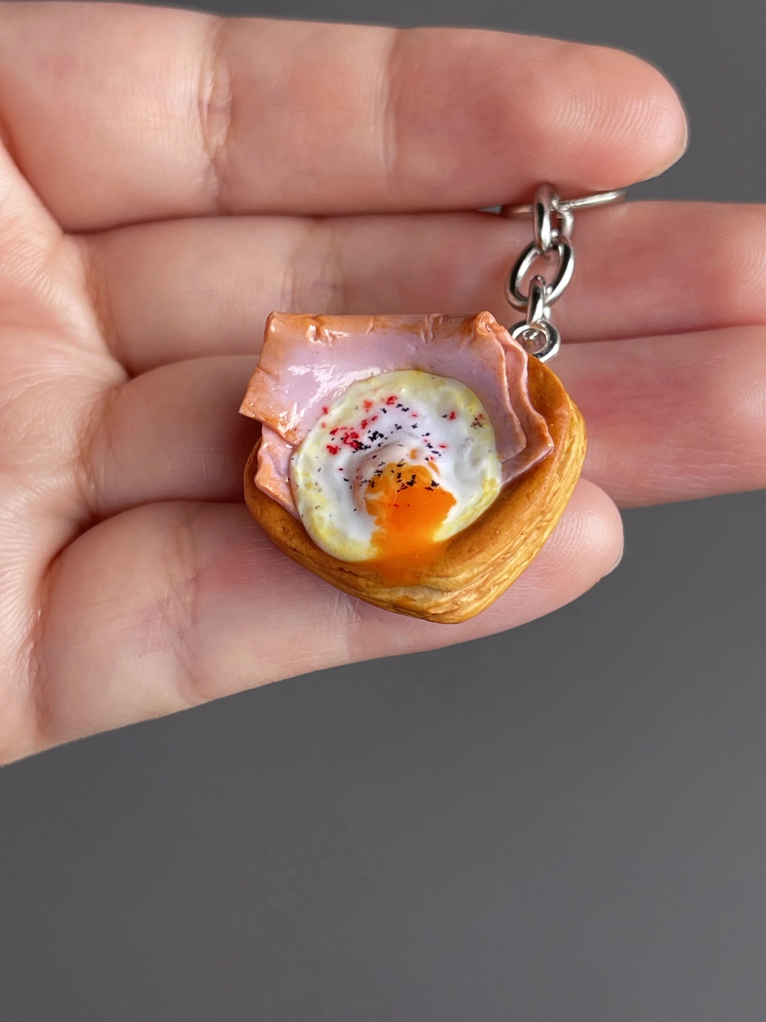 A hand holding a miniature polymer clay danish keychain with a piece of ham and egg with runny egg yolk.