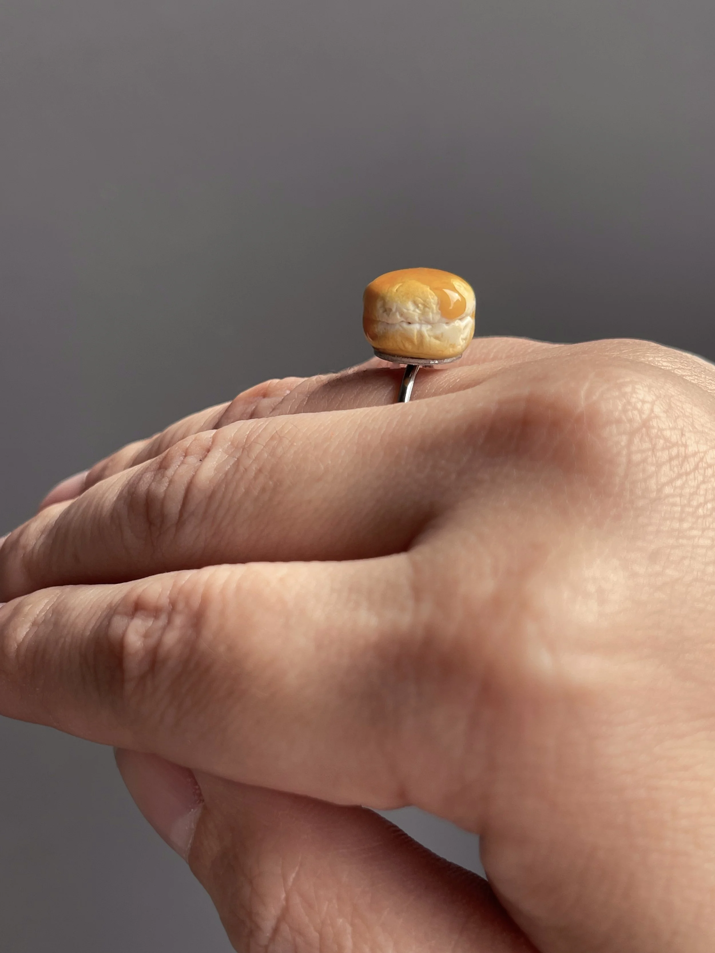 A hand wearing an adjustable ring topped with a miniature polymer clay biscuit with resin dripping honey.