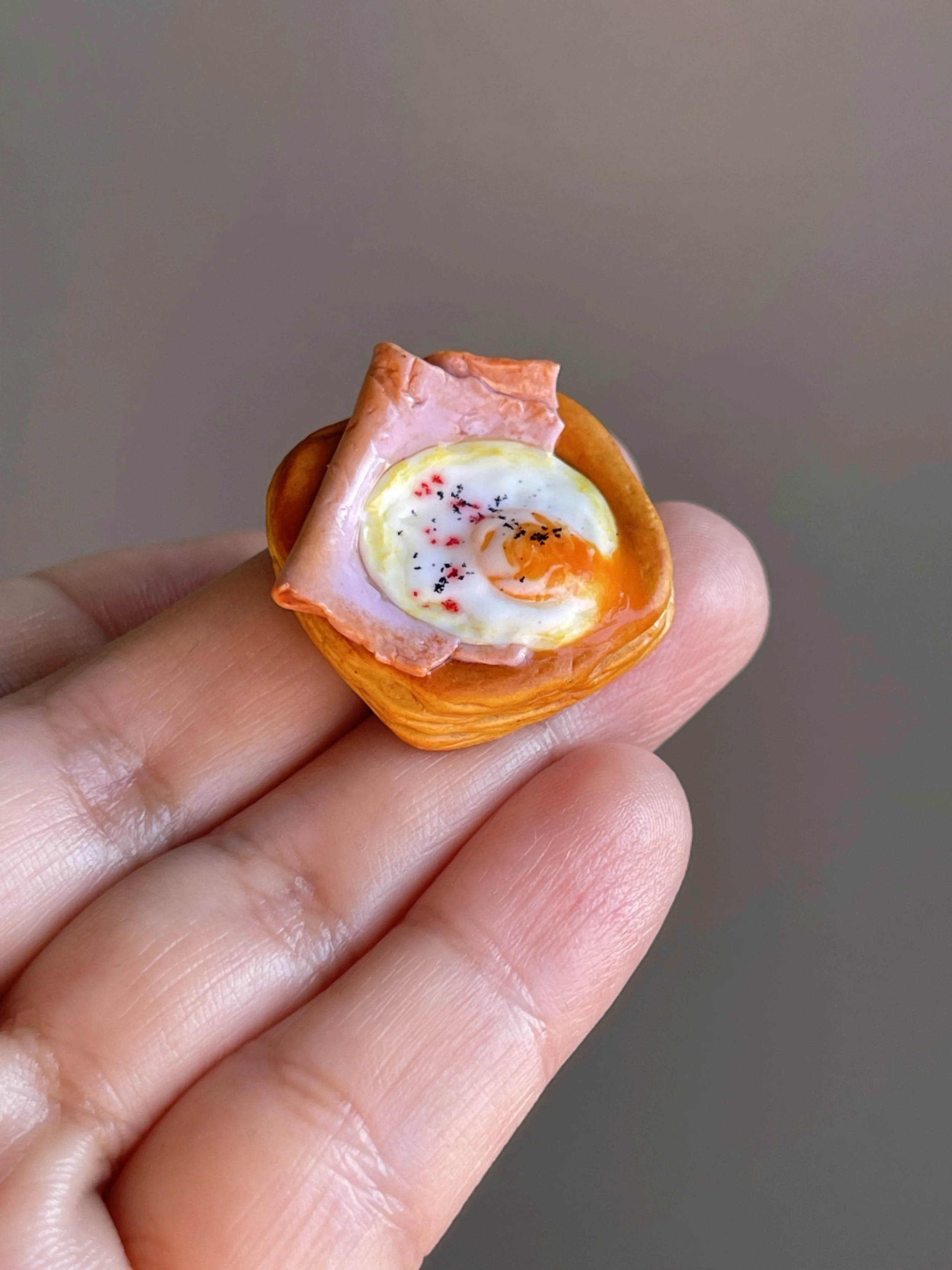 A hand holding a miniature realistic polymer clay ham and egg danish magnet.