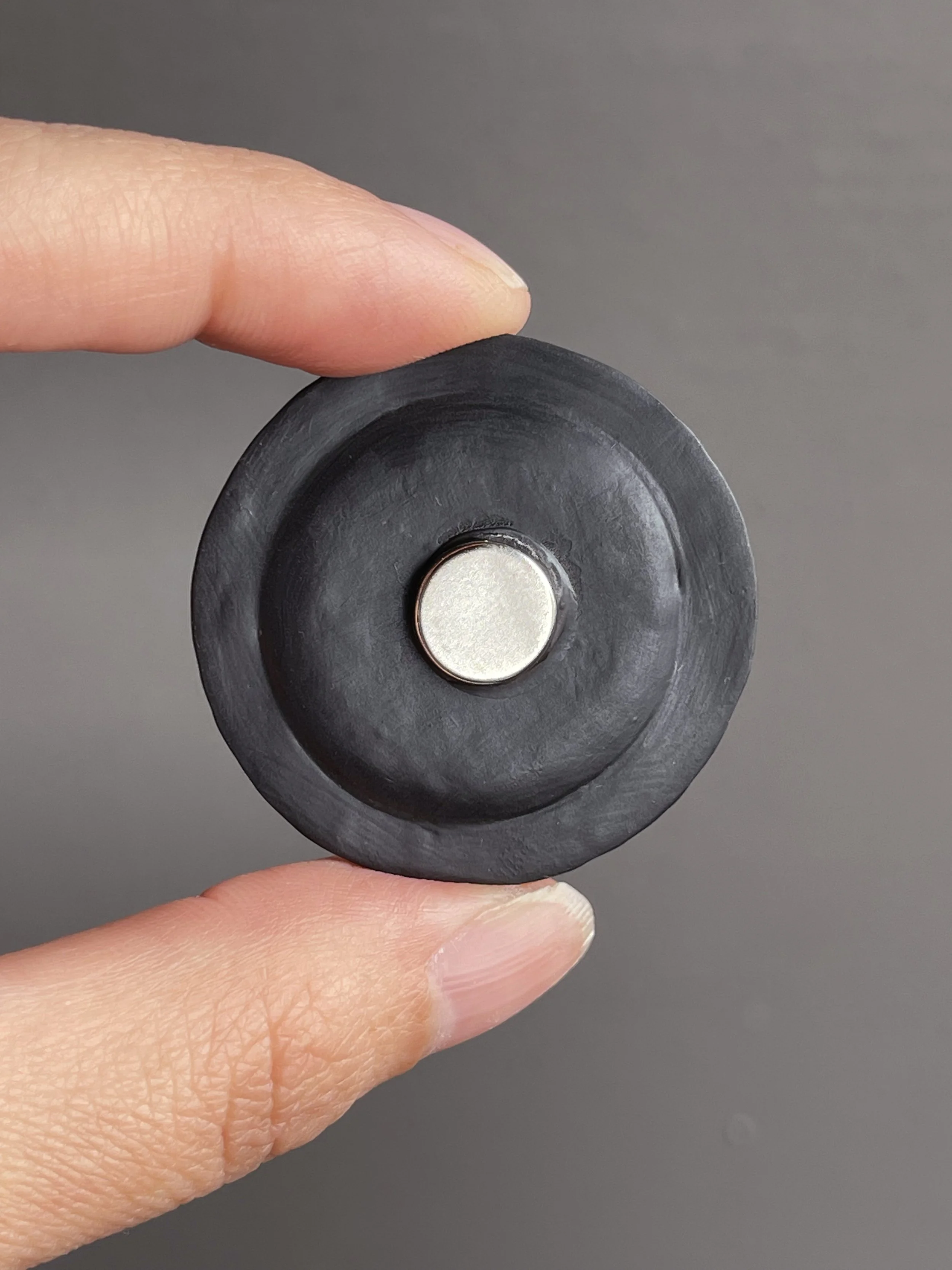 Fingers holding a miniature polymer clay kimchi fried rice magnet topped with a fried egg and green onions atop a black plate showing the back.