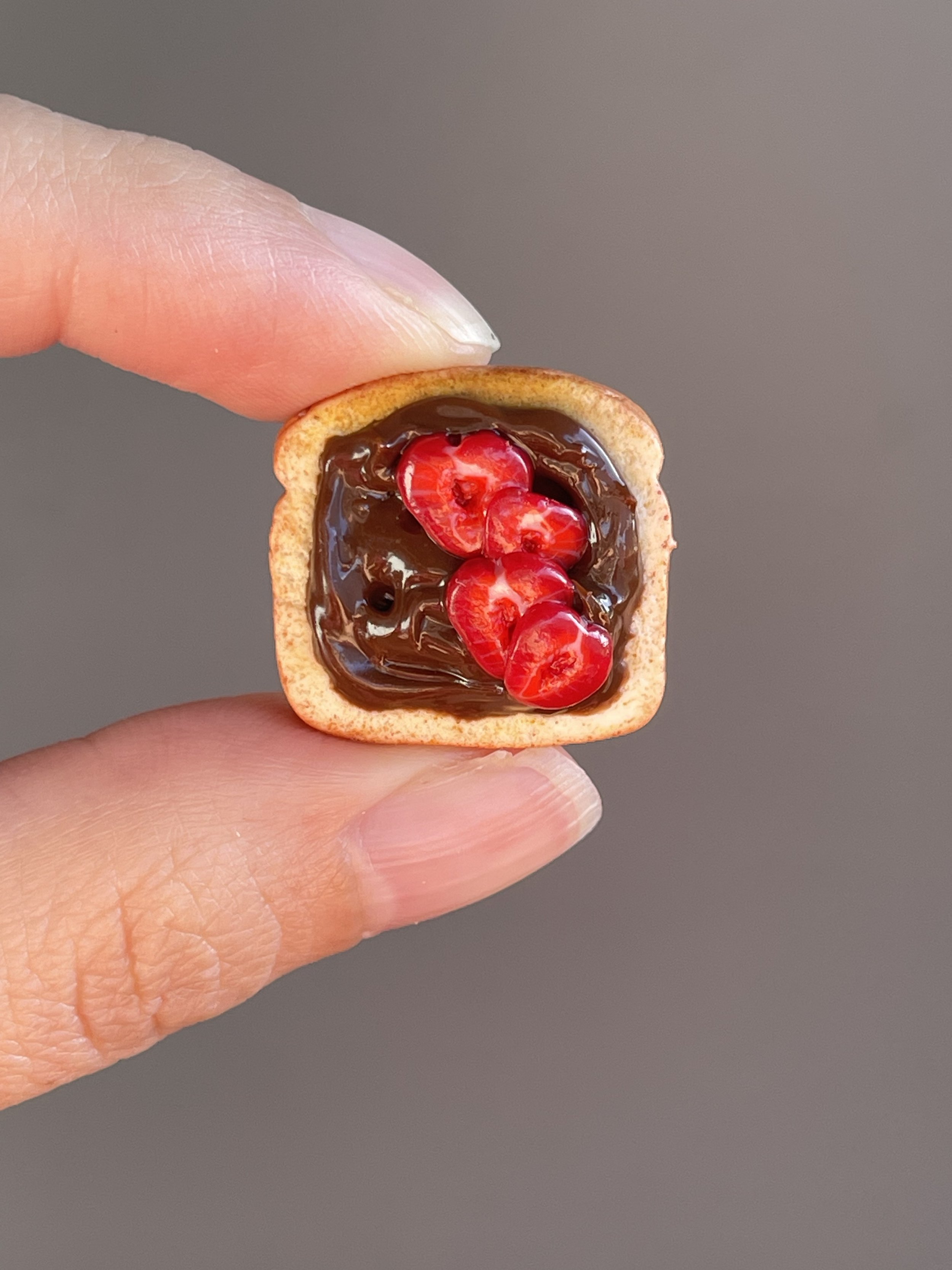 A hand holding a miniature polymer clay hazelnut spread and sliced strawberries toast magnet.