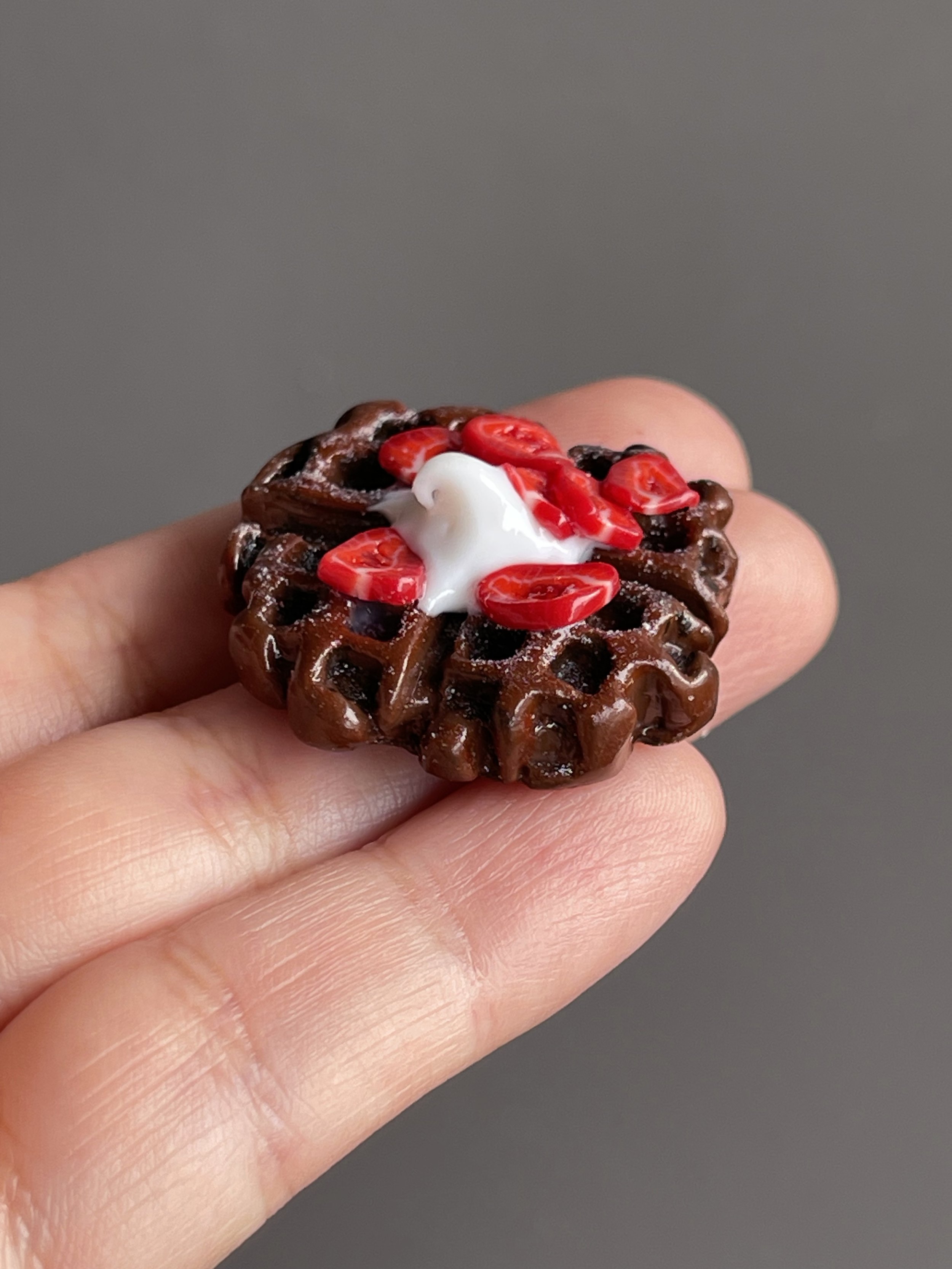 A hand holding a miniature polymer clay chocolate waffle magnet topped with strawberries and whipped cream.