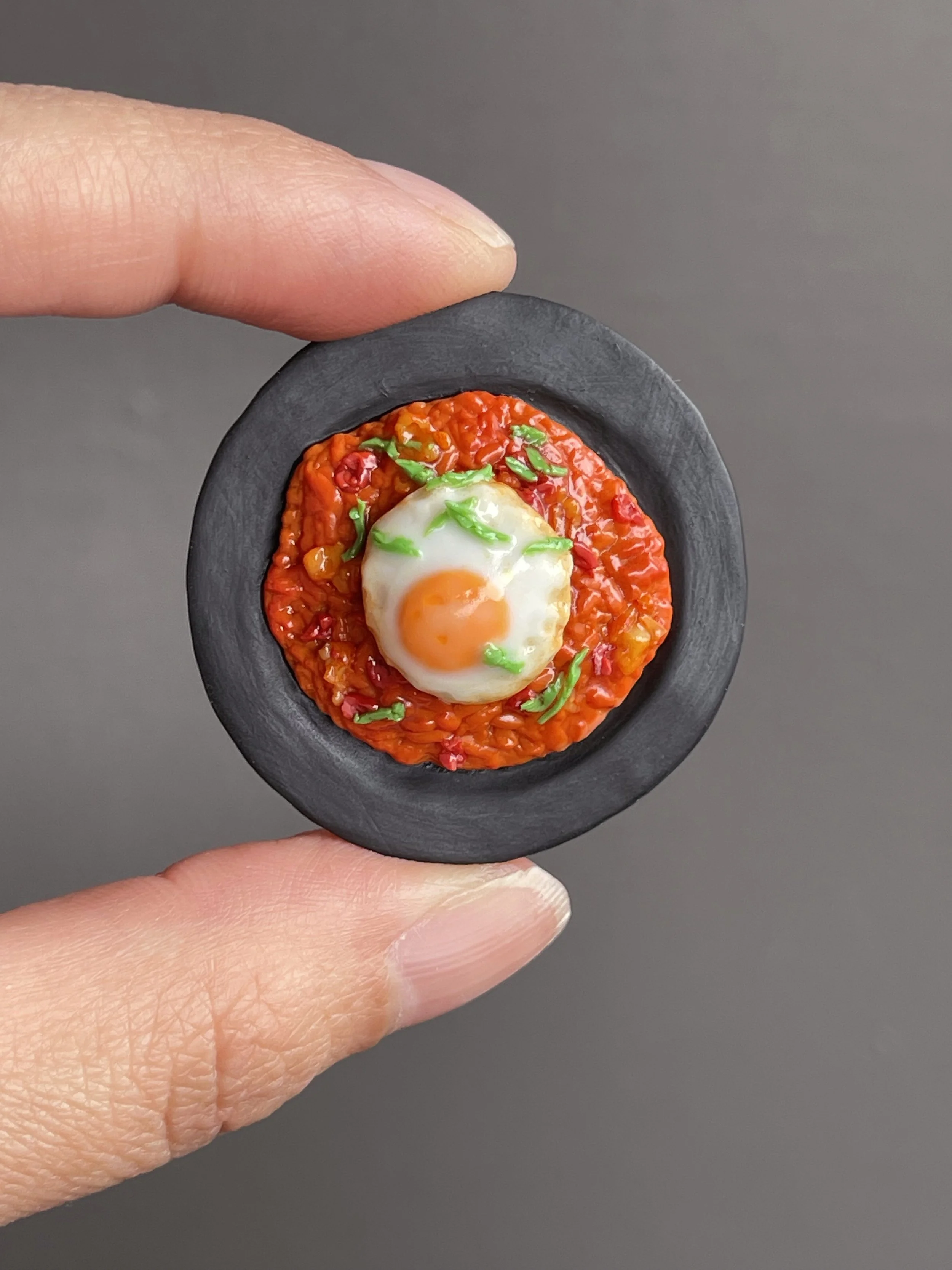 Fingers holding a miniature polymer clay kimchi fried rice magnet topped with a fried egg and green onions atop a black plate.