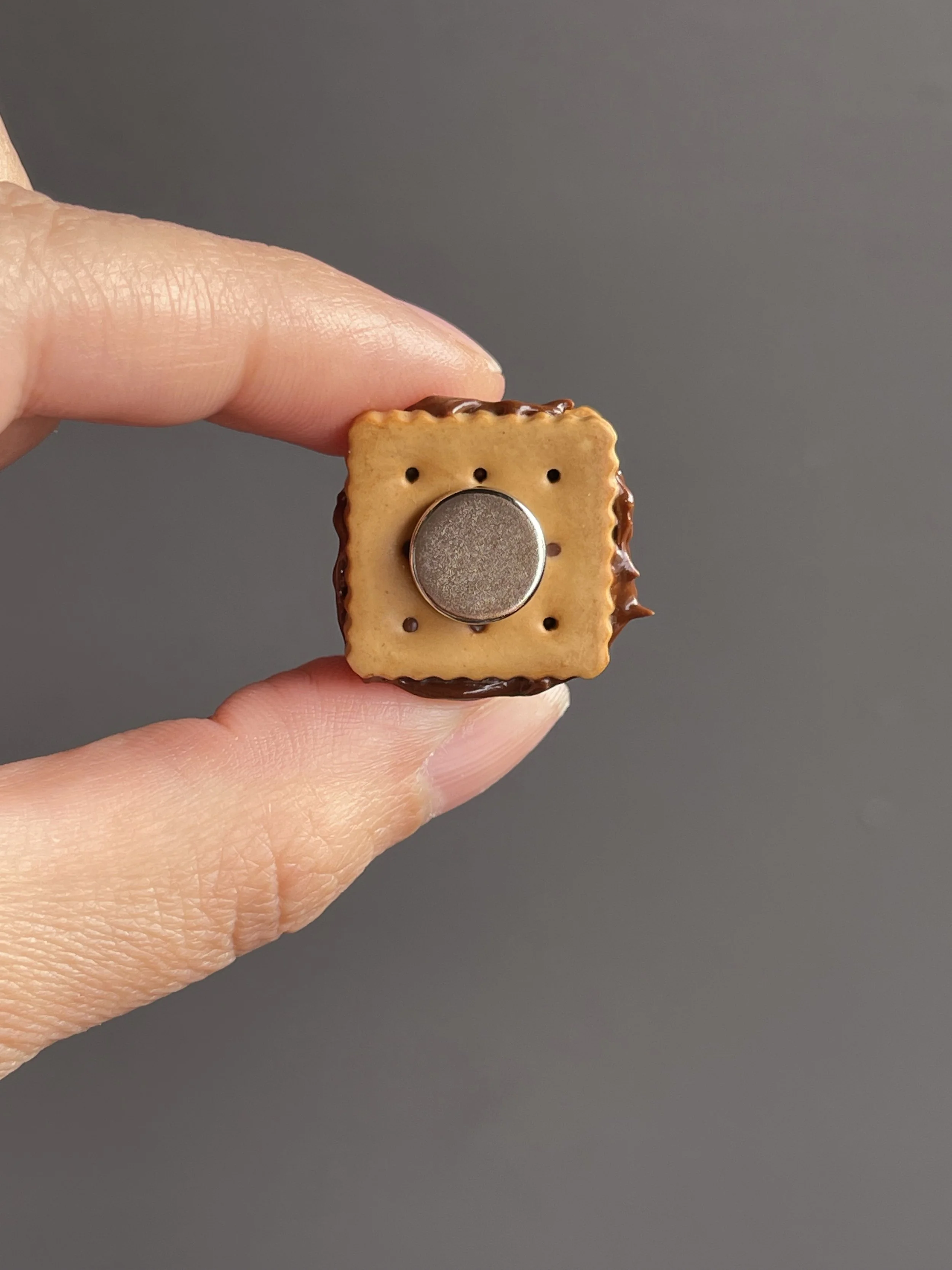 Fingers holding a miniature polymer clay s’more magnet showing the back.