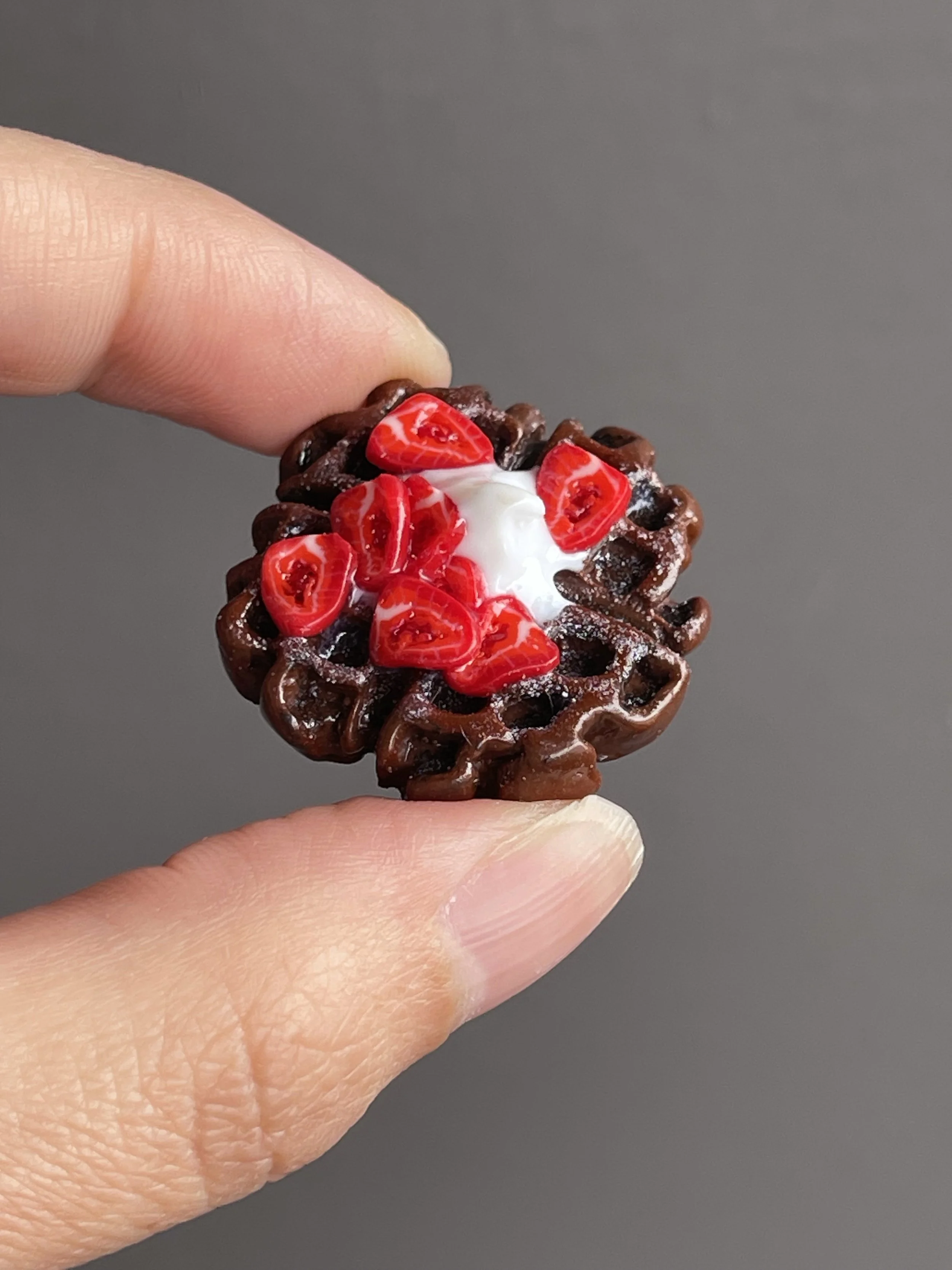 Fingers holding a miniature polymer clay chocolate waffle magnet topped with strawberries and whipped cream.