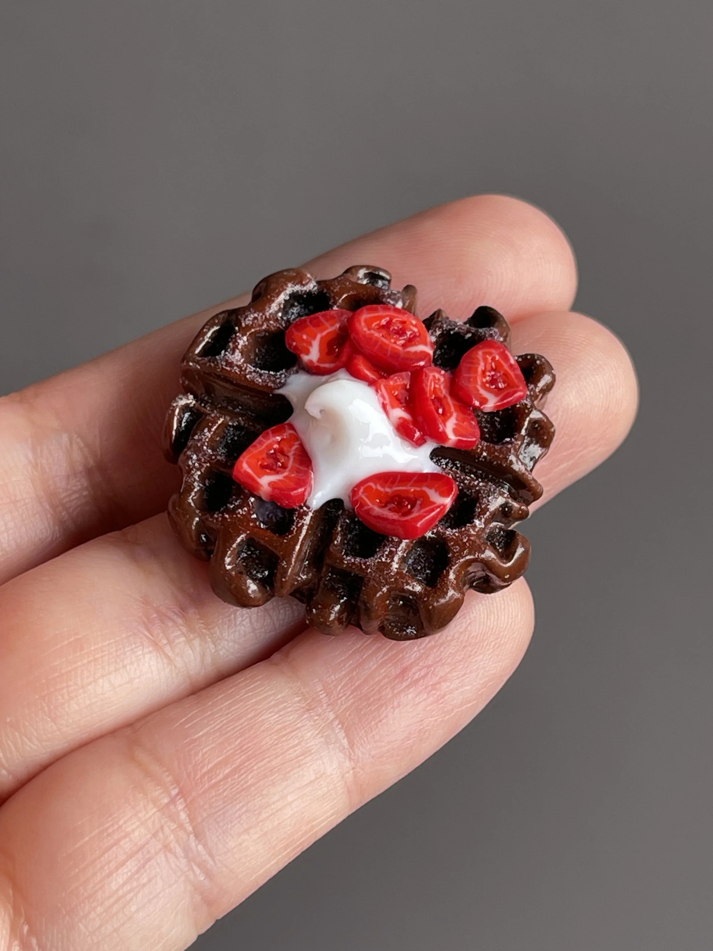 A hand holding a miniature polymer clay chocolate waffle magnet topped with strawberries and whipped cream.