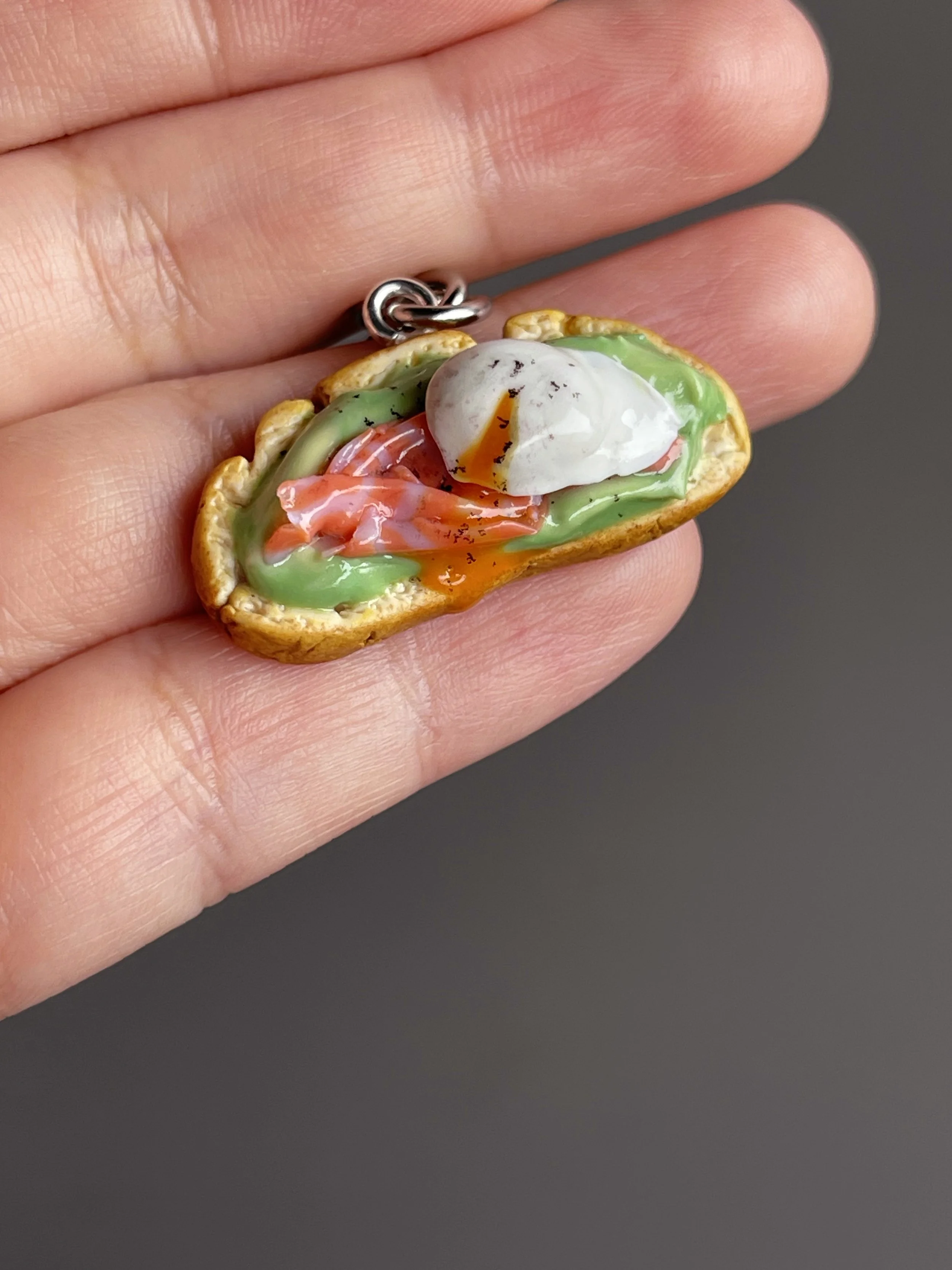 A hand holding a miniature polymer clay avocado toast keychain with smoked salmon, avocado mash, and a runny poached egg.