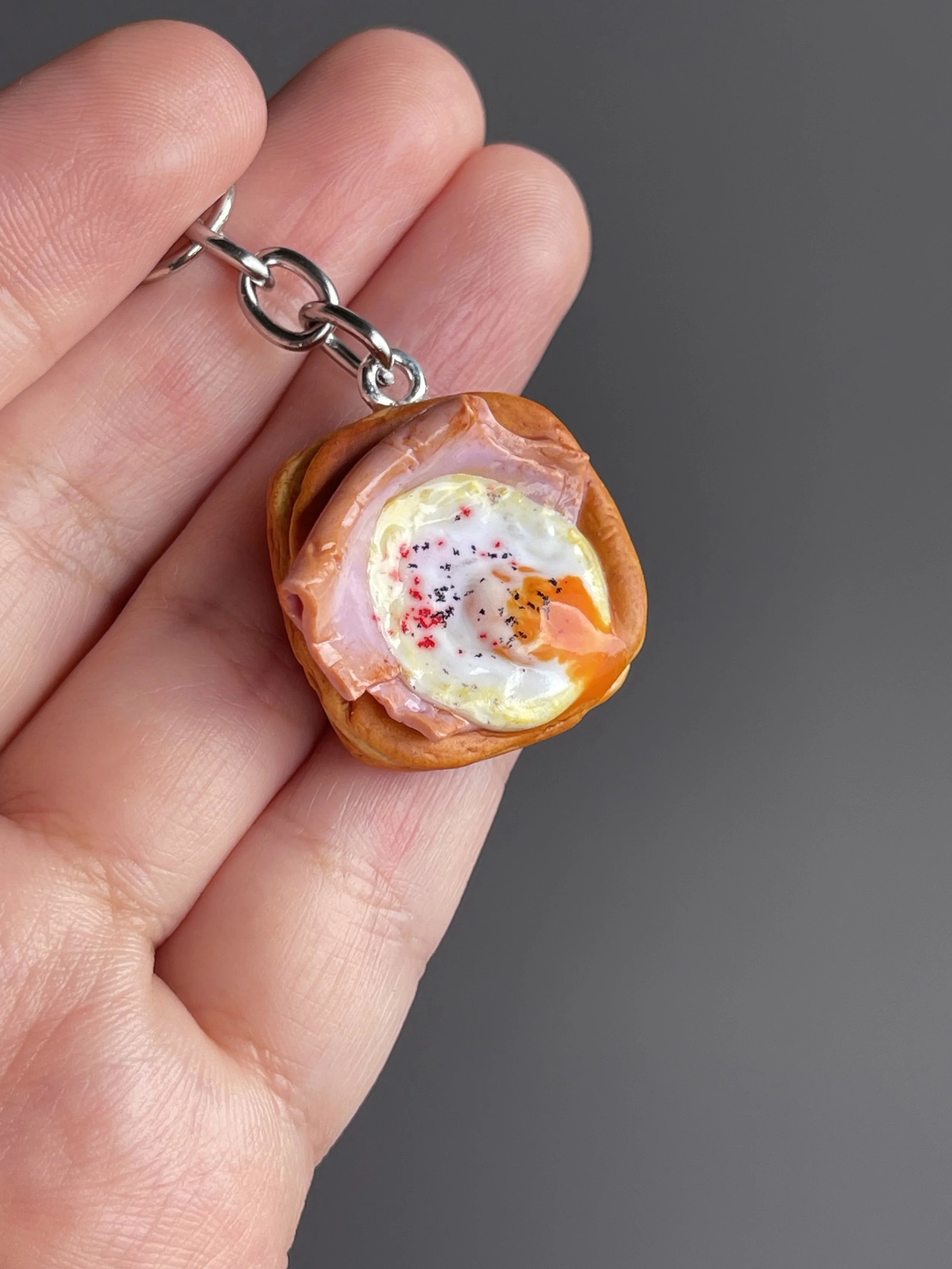 A hand holding a miniature polymer clay danish keychain with a piece of ham and egg with runny egg yolk.