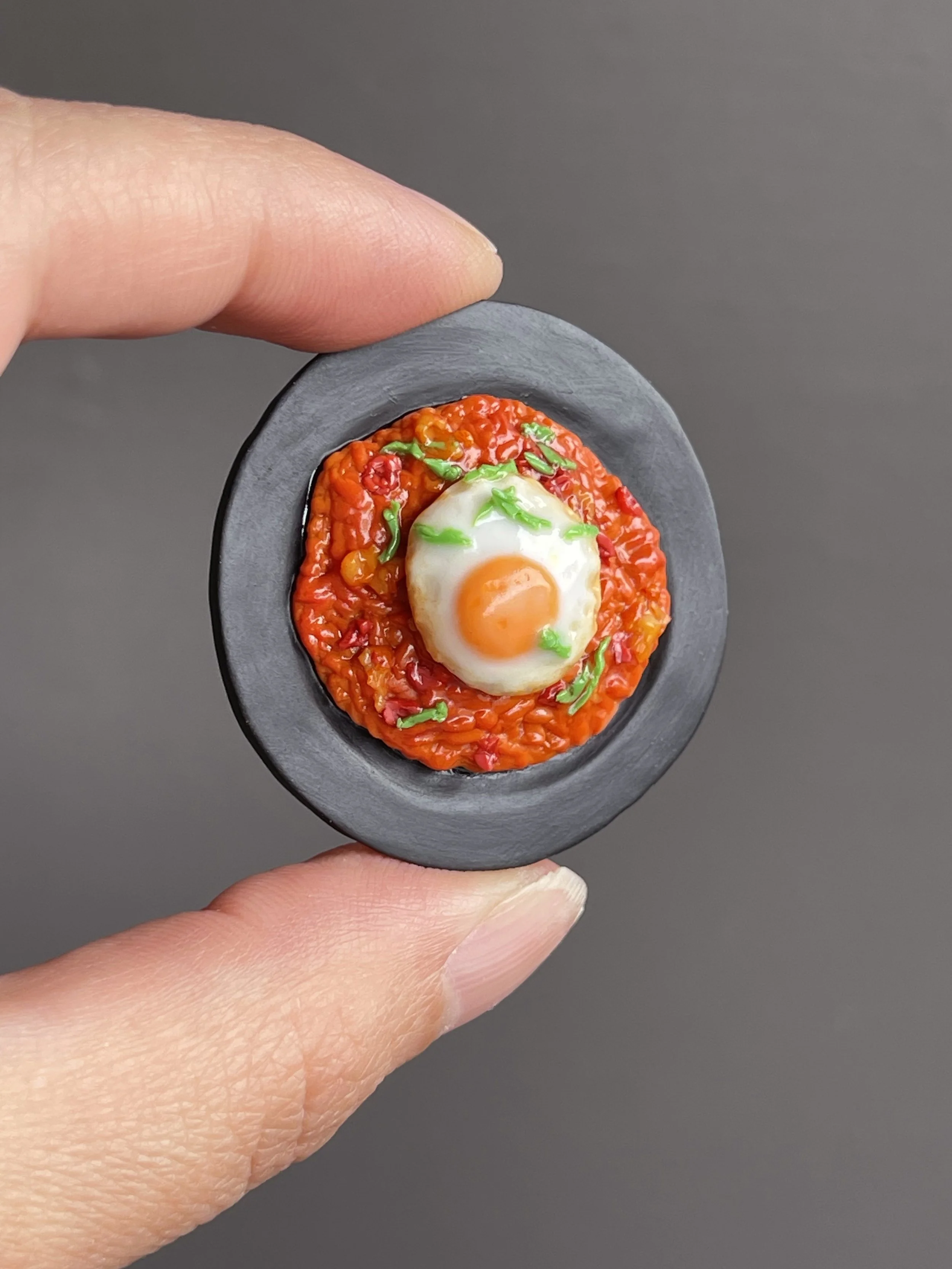 Fingers holding a miniature polymer clay kimchi fried rice magnet topped with a fried egg and green onions atop a black plate.