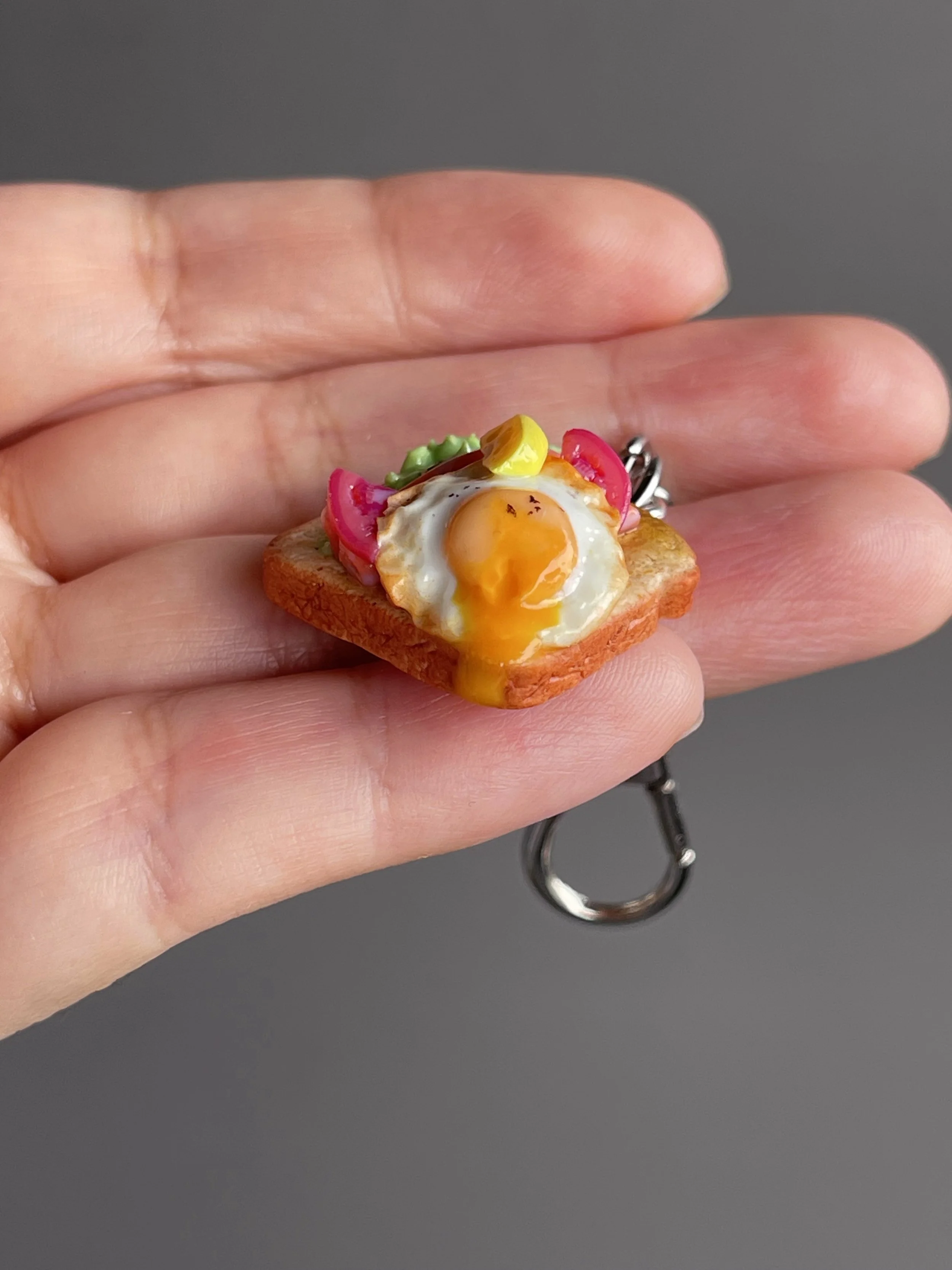 A hand holding a miniature polymer clay avocado, smoked salmon, tomato toast keychain with runny egg yolk and lemon slice garnish.