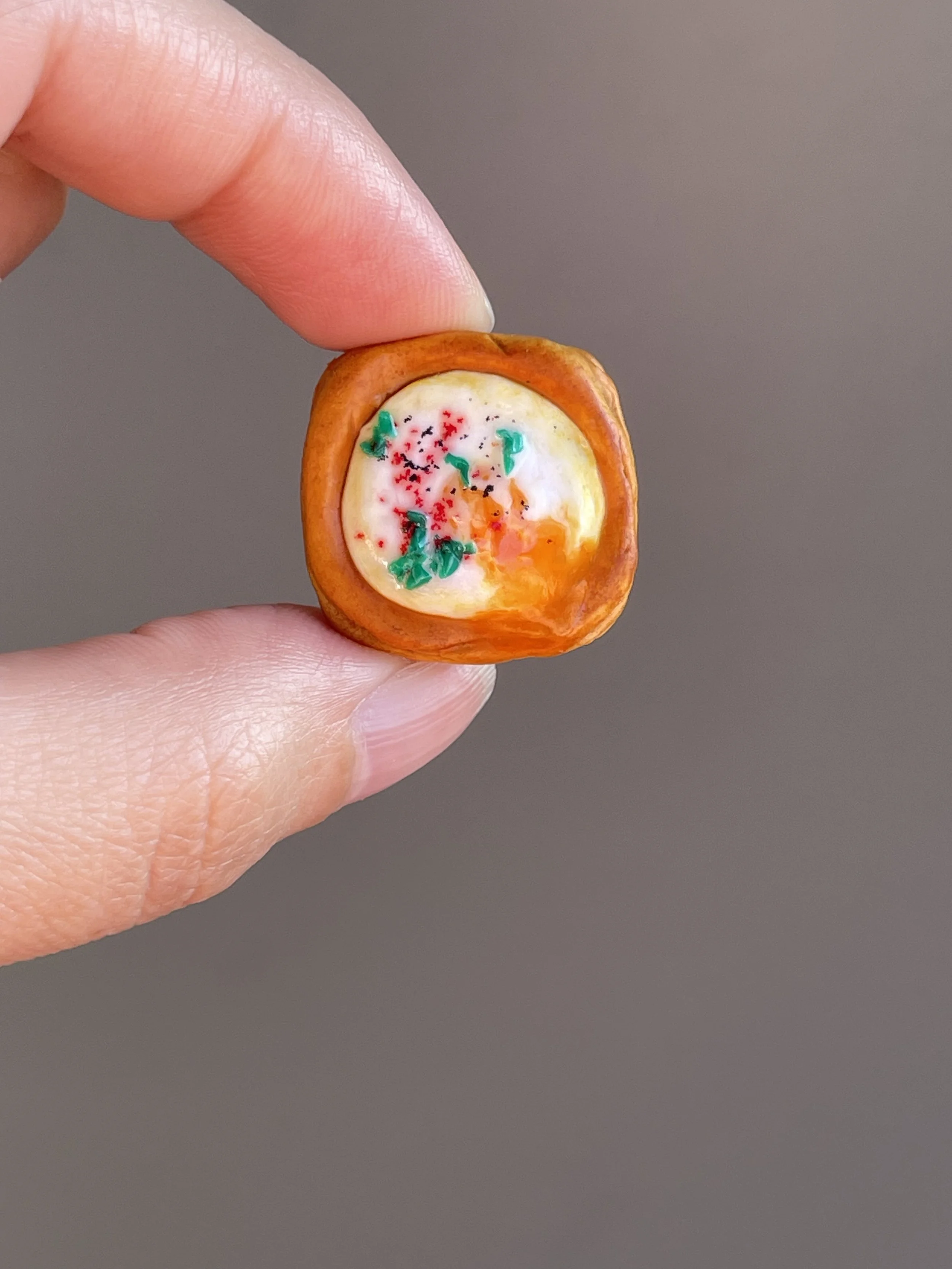 Fingers holding a miniature polymer clay egg danish magnet with runny egg yolk.