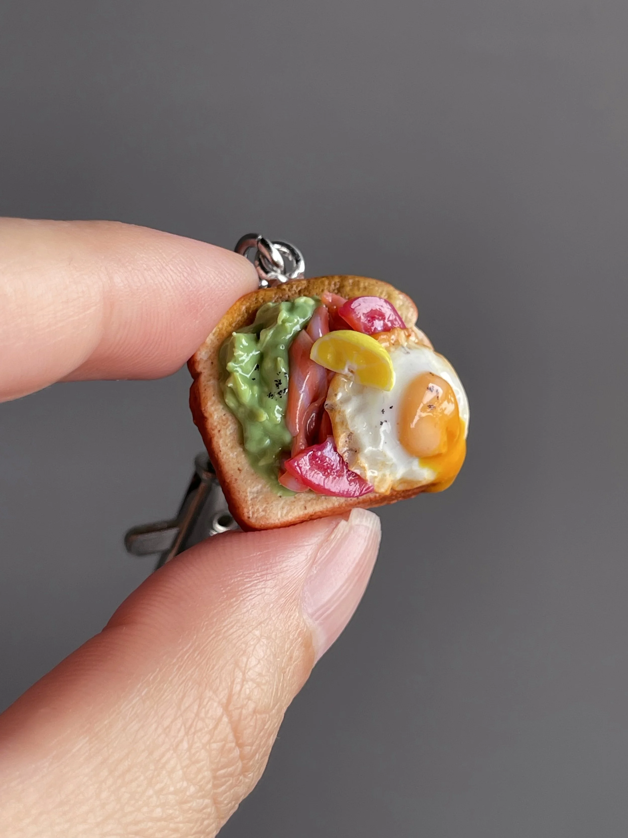 A hand holding a miniature polymer clay avocado, smoked salmon, tomato toast keychain with runny egg yolk and lemon slice garnish.