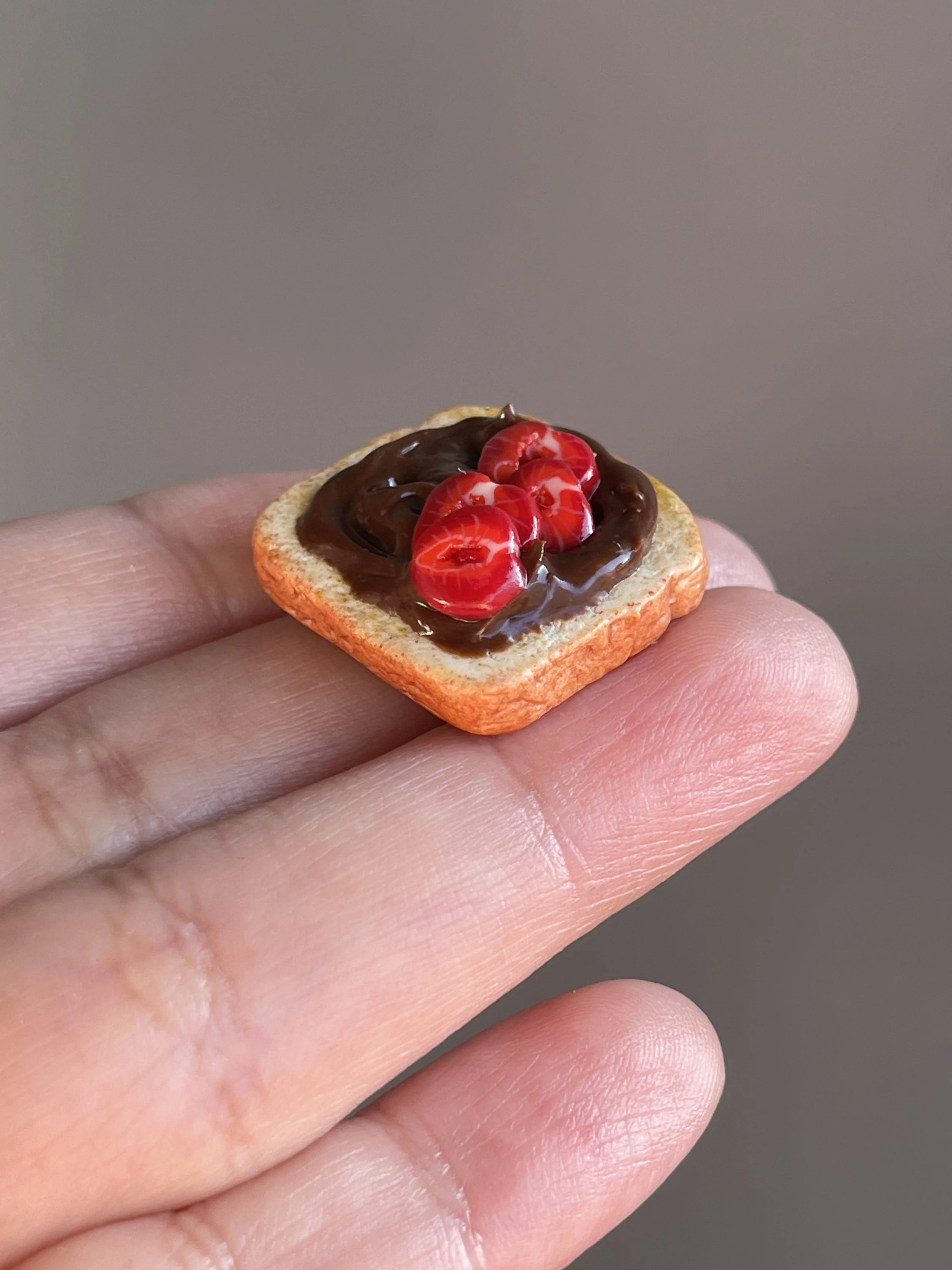 A hand holding a miniature polymer clay hazelnut spread and sliced strawberries toast magnet.