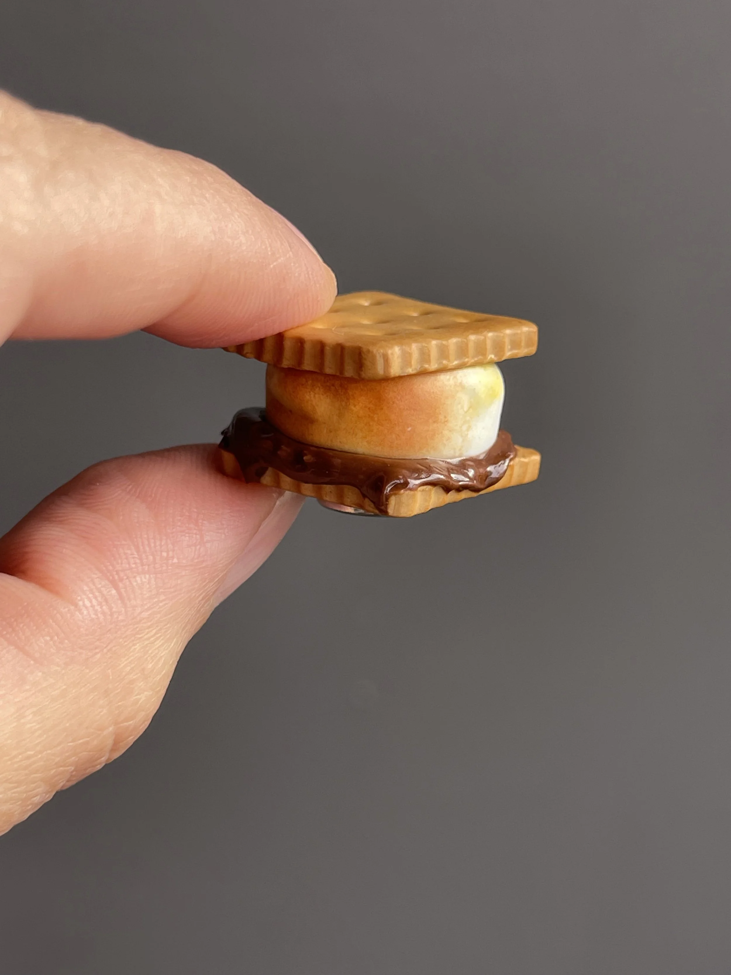 Fingers holding a miniature polymer clay s’more magnet.