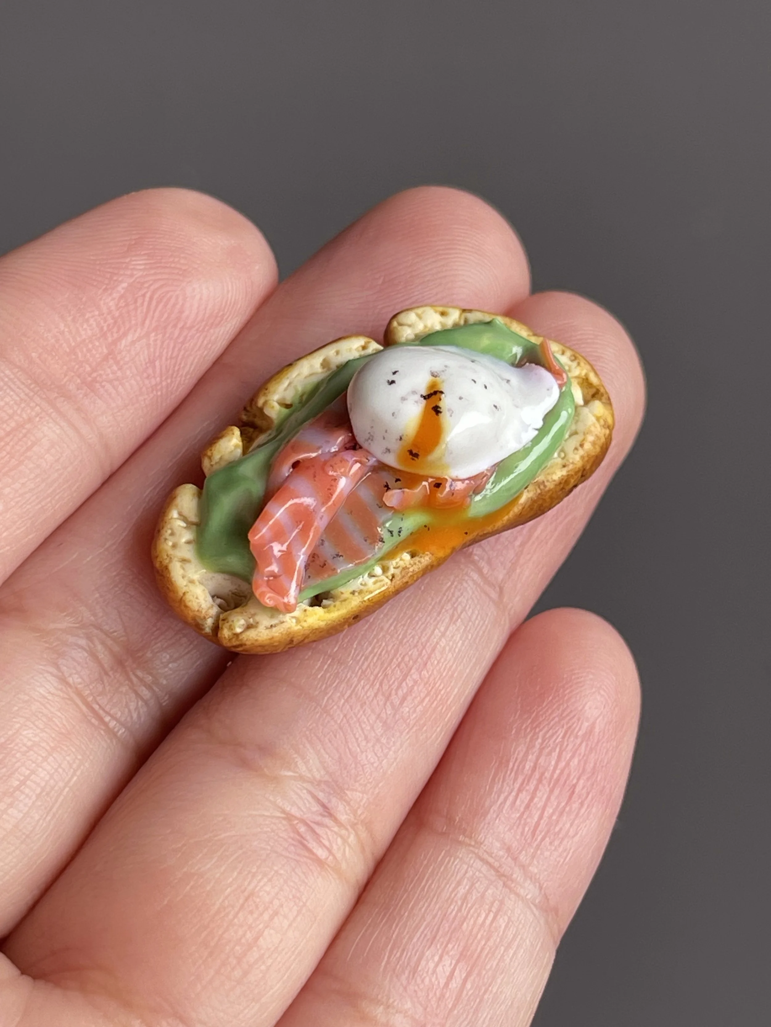 A hand holding a miniature polymer clay avocado toast magnet with smoked salmon, avocado mash, and a runny poached egg.