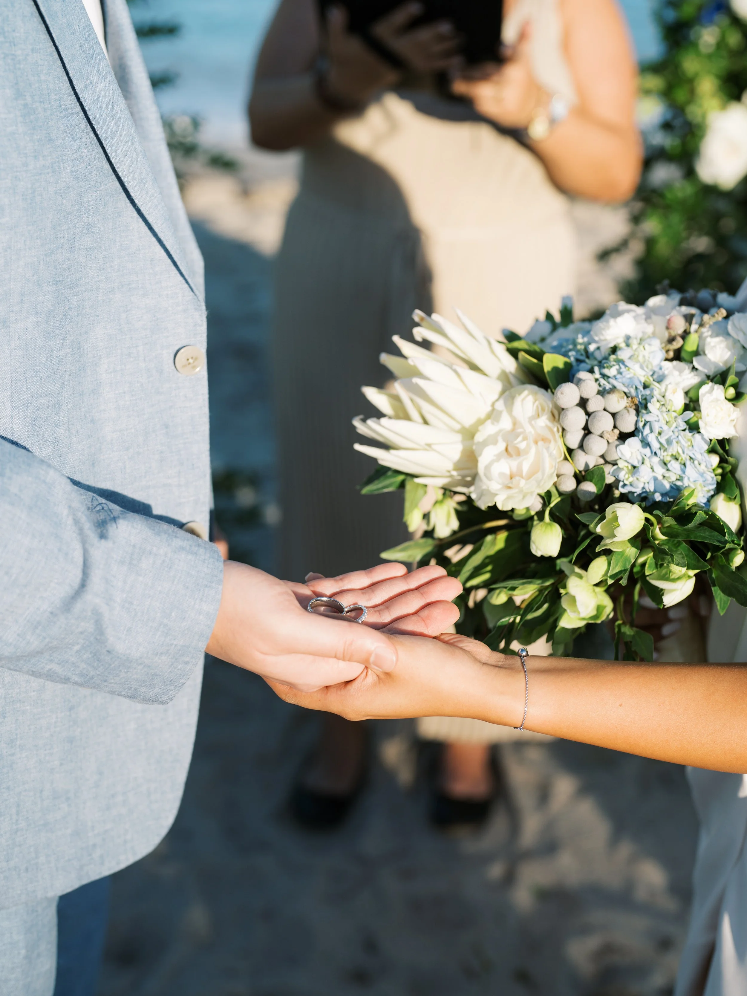 A couple exchanging rings during a wedding ceremony on a beach, with a bouquet of white and blue flowers in the background.
