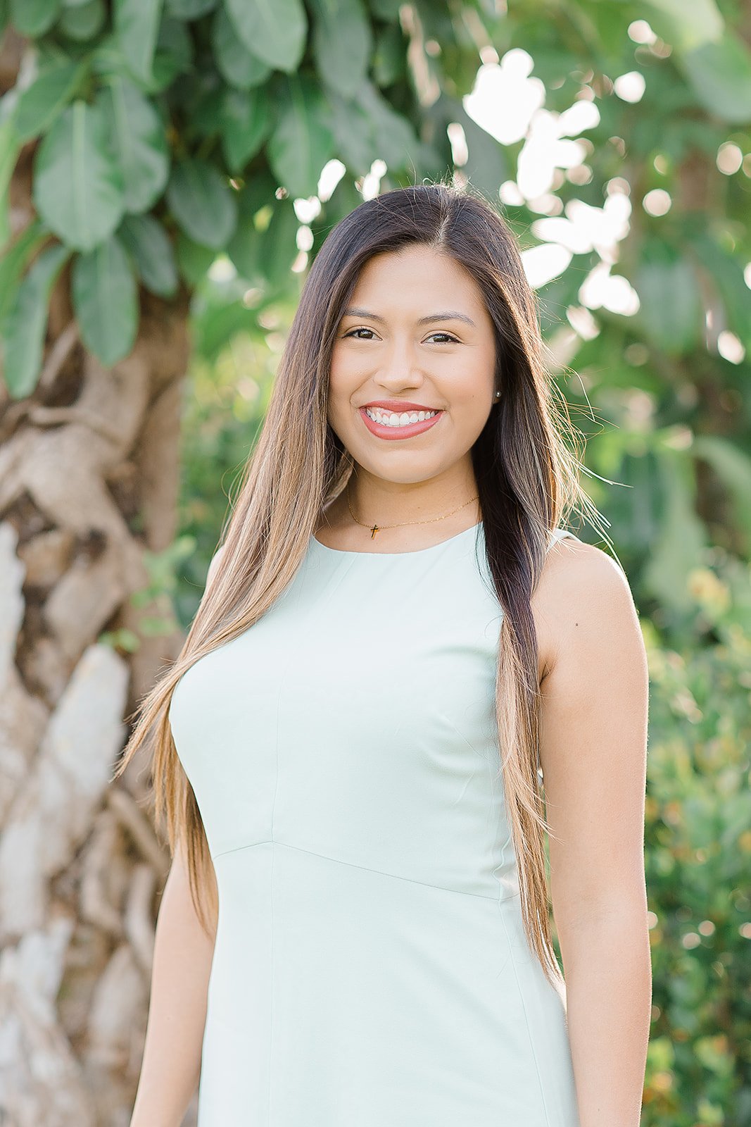 A young woman with long, straight hair wearing a white sleeveless dress, smiling outdoors with a green leafy background.