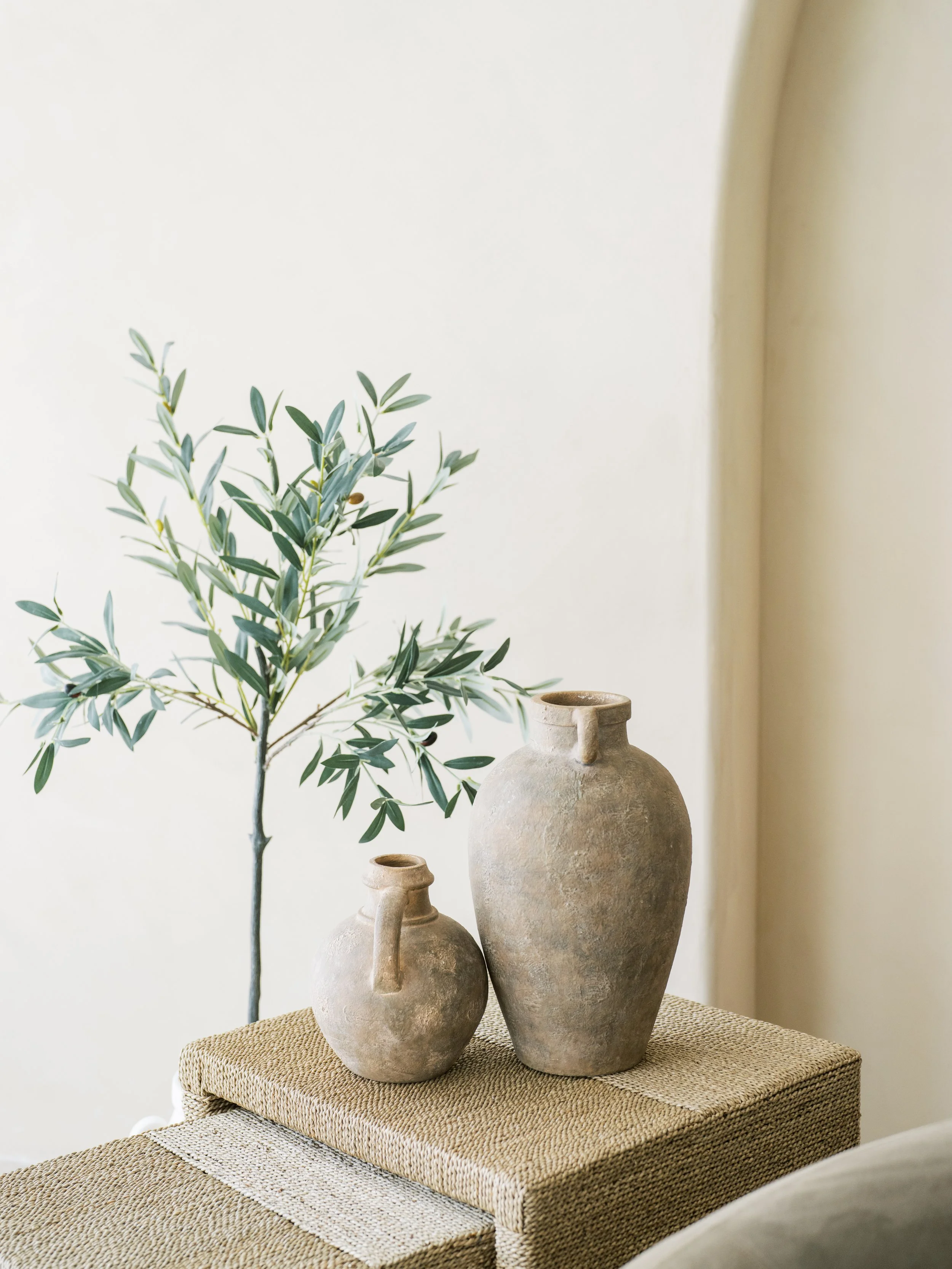 Two textured ceramic vases, one taller and one smaller, placed on a woven surface with a green leafy plant in the background.