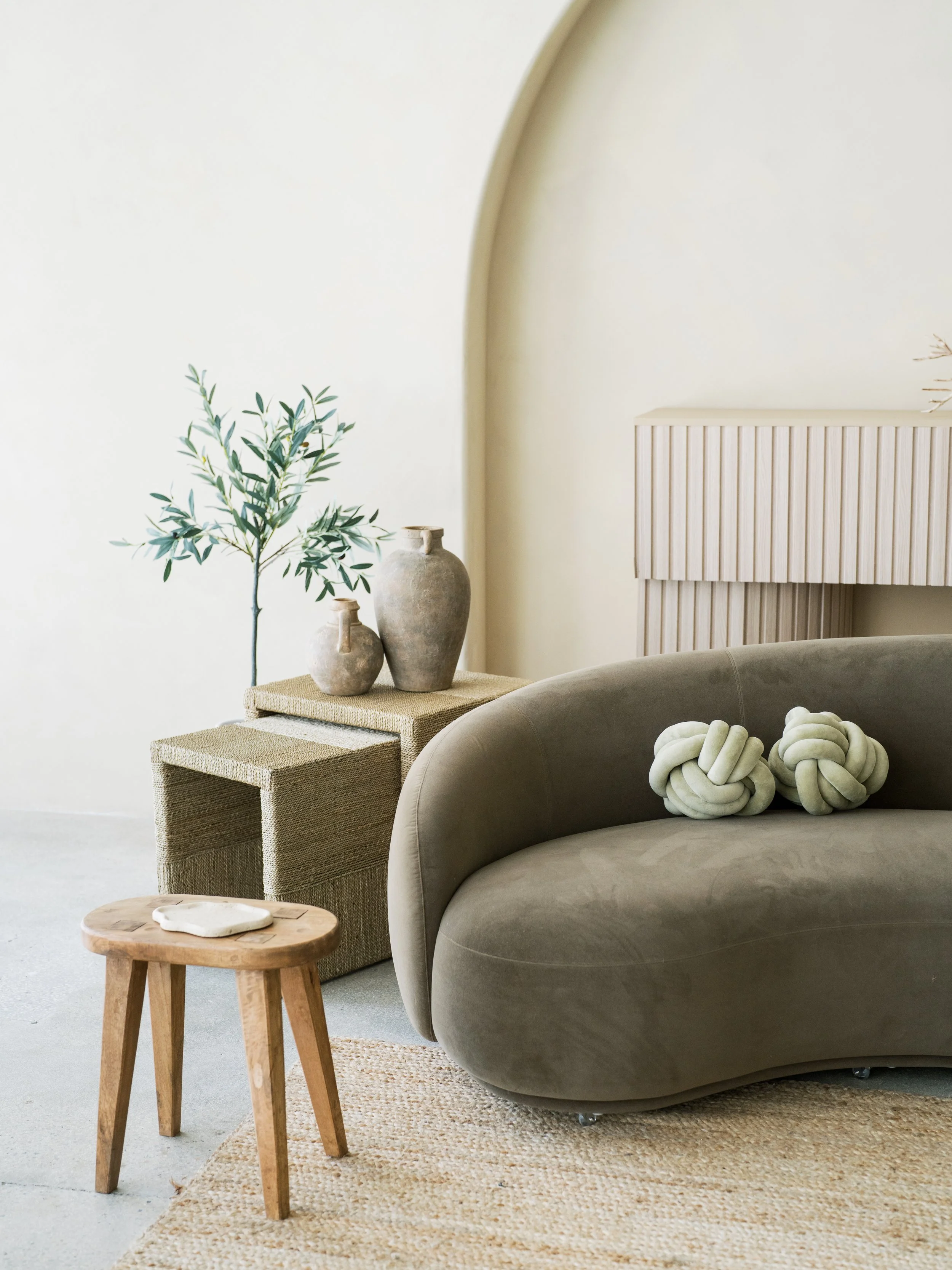 Minimalist living room with beige wall, arched corner, beige sideboard, gray plush sofa with two knotted decorative pillows, small wooden stool with a white plate, textured wicker table with three ceramic vases, and a potted olive tree.