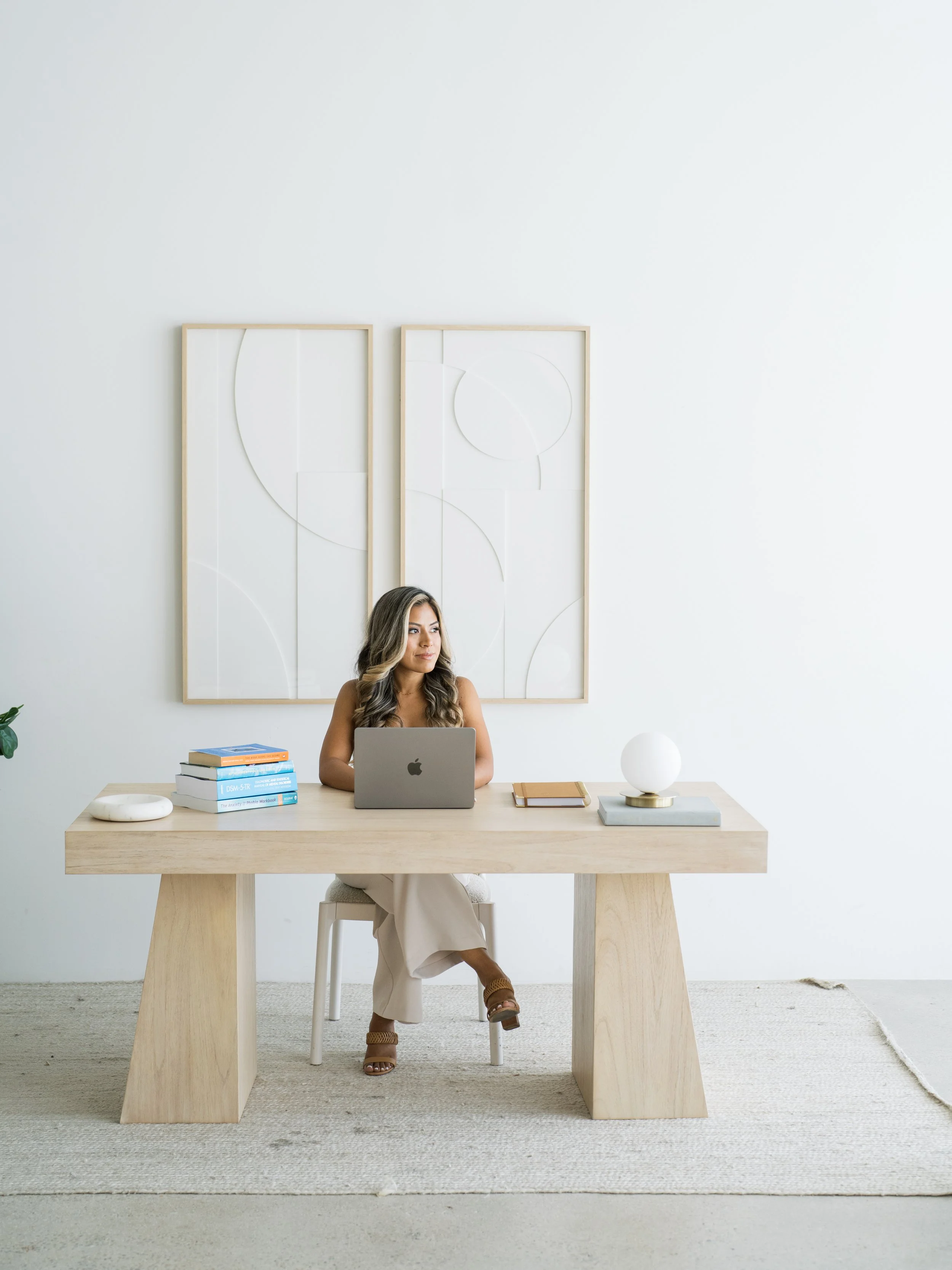 A woman sitting at a light wooden desk with a laptop, books, and decor in a minimalistic, white room.