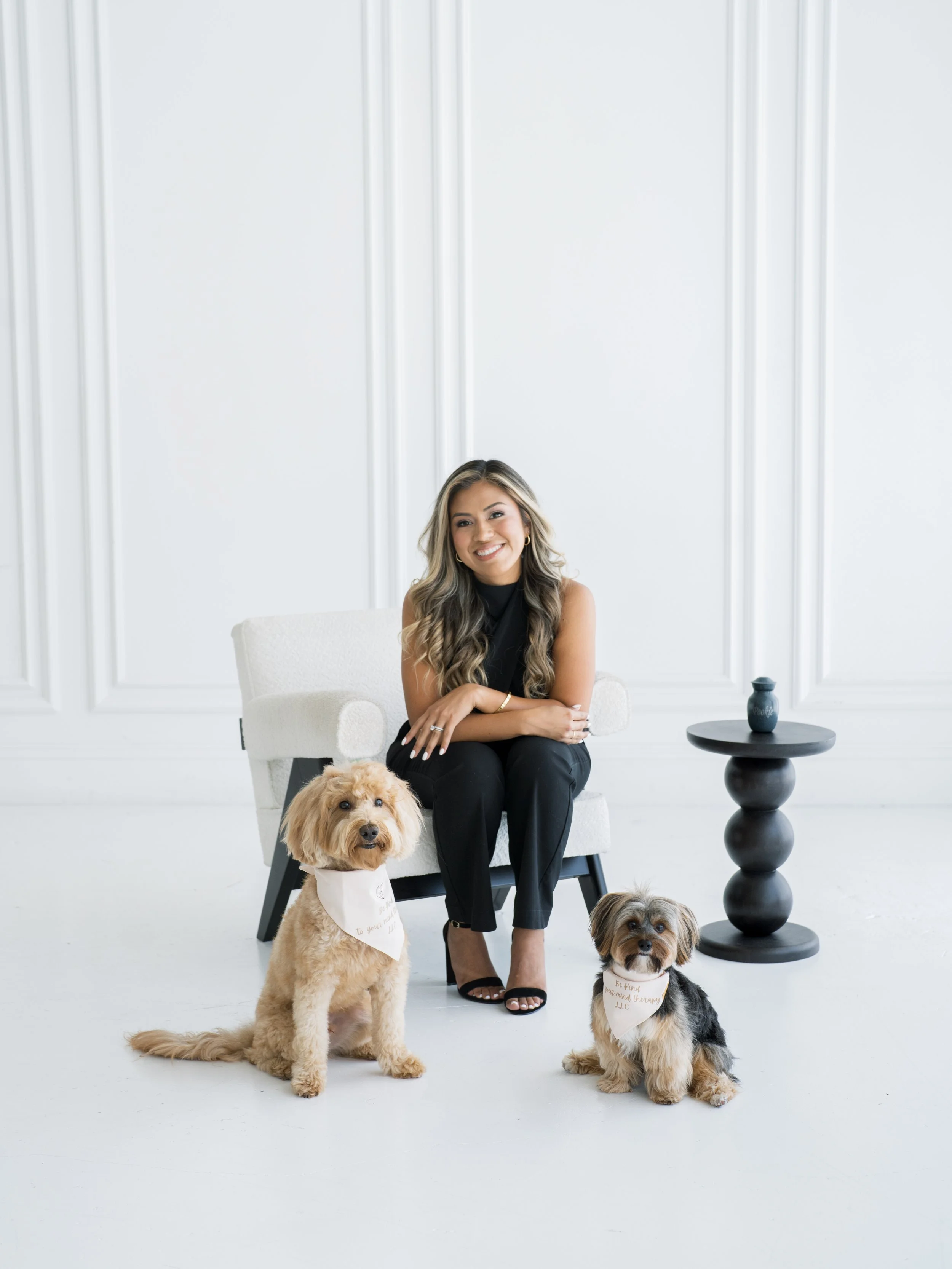 A woman sitting on a white armchair with two small dogs, one sitting and one lying on the floor, in a bright minimalist room with white walls, a black side table, and a small blue decorative object.