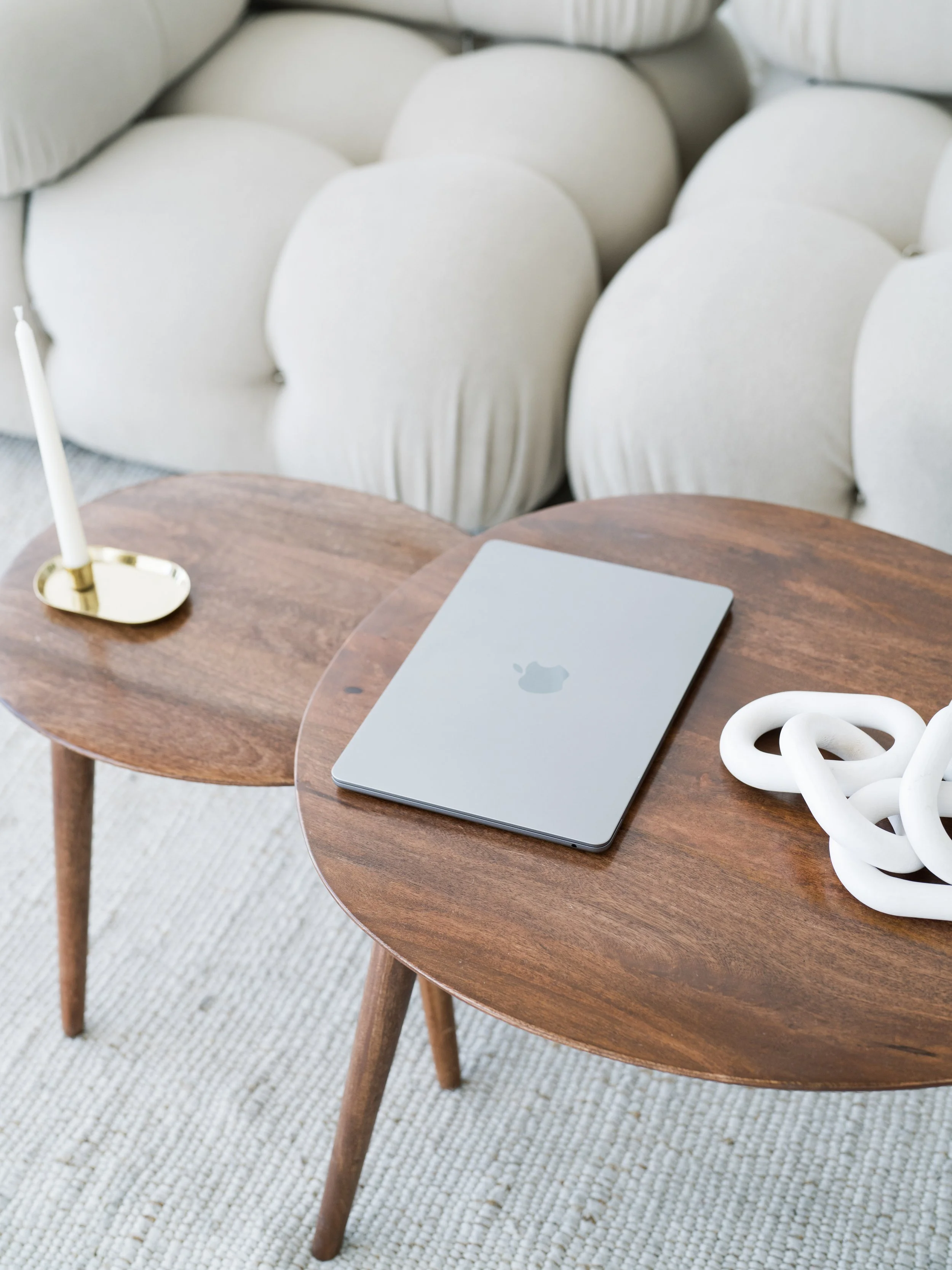 Two wooden side tables, one with a closed silver MacBook and a white decorative sculpture, and the other with a white candlestick holder, positioned in front of a cream-colored sofa with plush, rounded cushions.