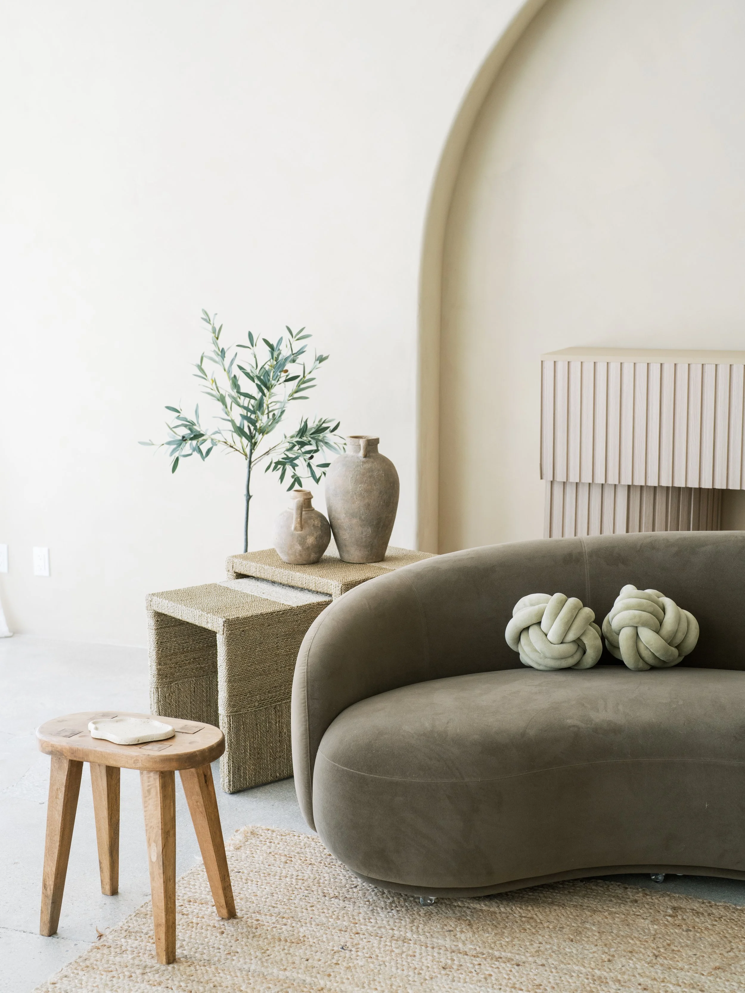 Minimalist living room with beige and taupe tones, featuring a curved taupe sofa with decorative knot cushions, a small wooden stool, a woven side table with ceramic vases, a tall potted plant, a textured beige wall, and a soft beige area rug.