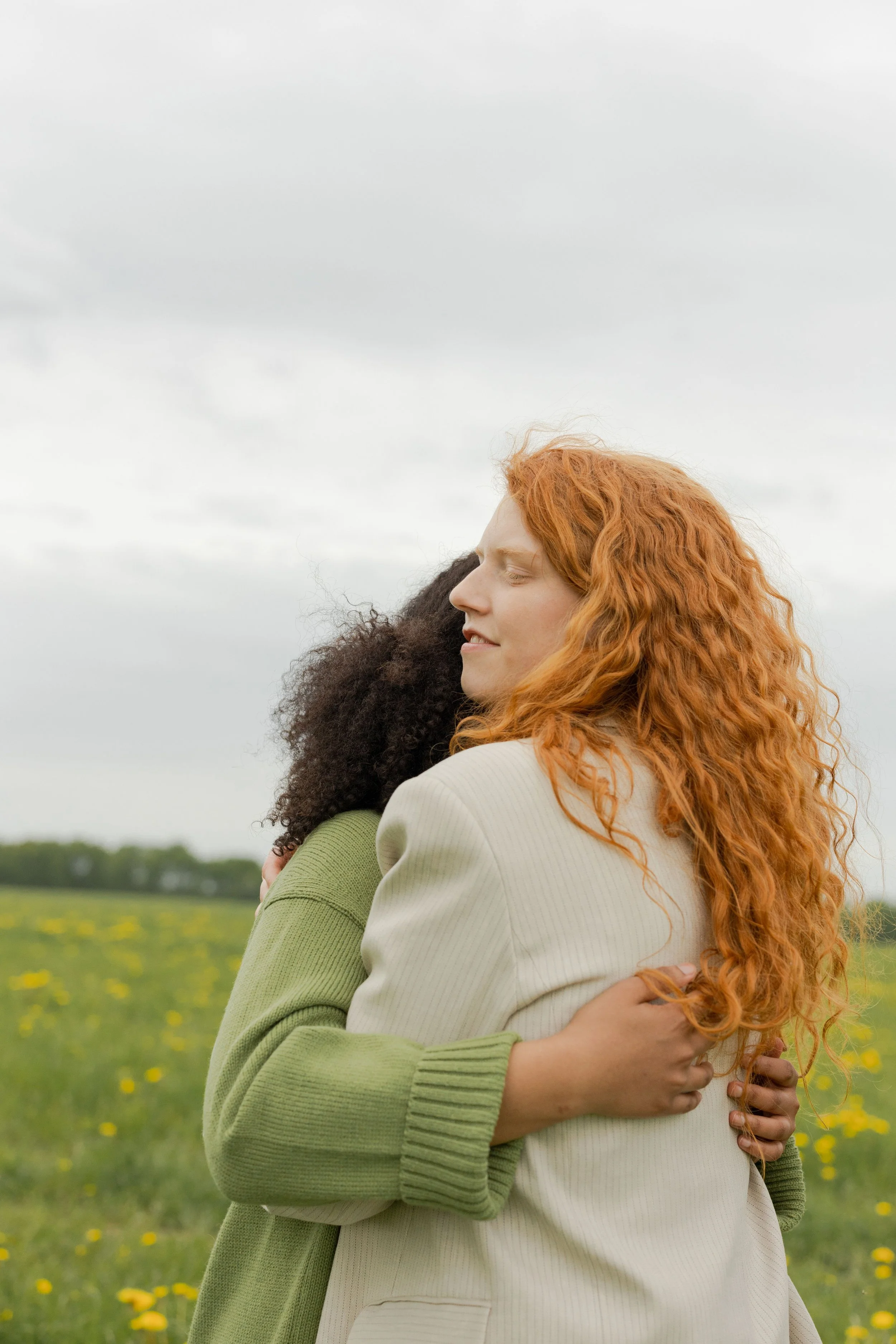 Zwei Menschen umarmen sich auf einer Blumenwiese, eine Person mit roten Locken und grüner Strickjacke, als Symbol für Halt und Unterstützung.