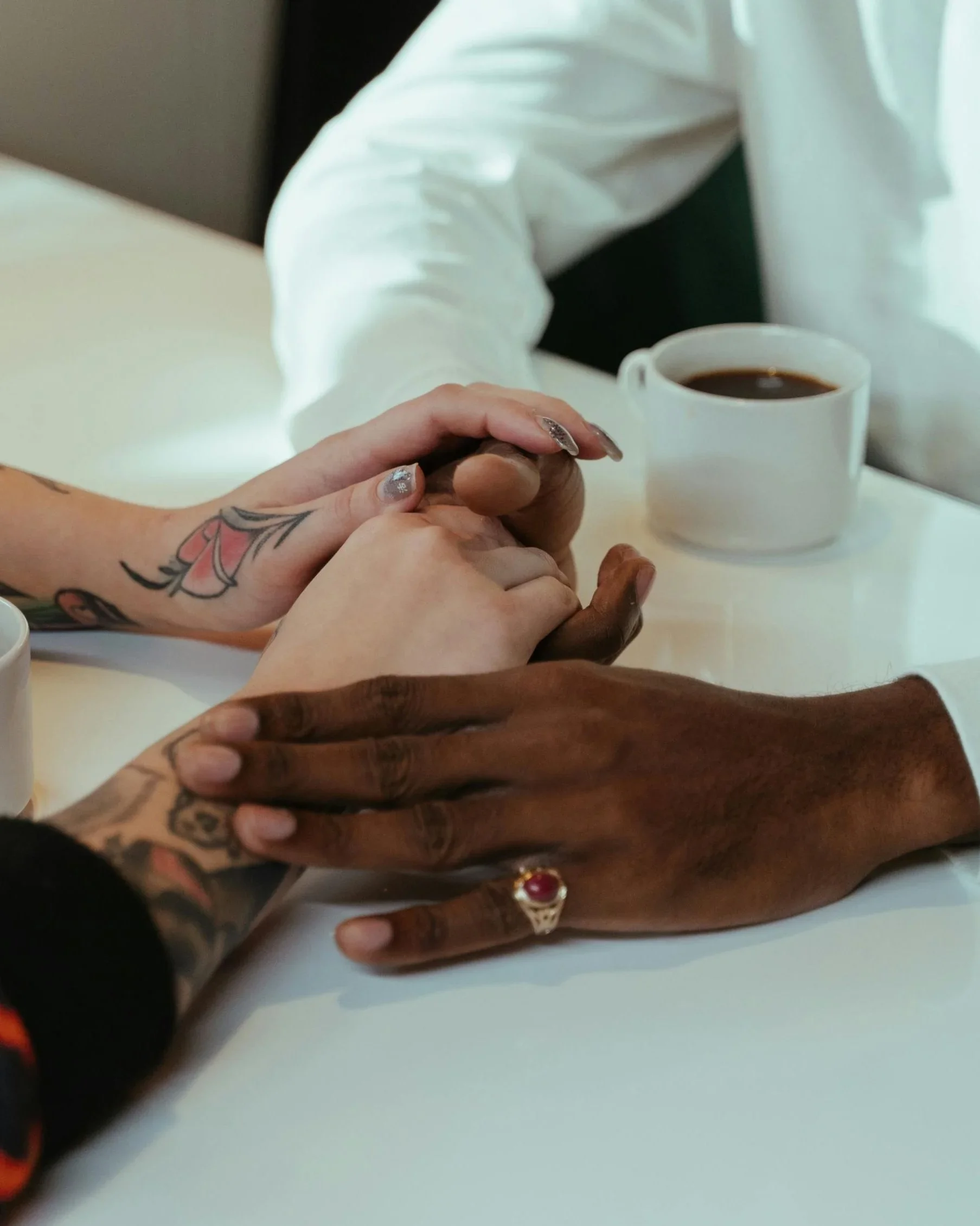 Mehrere Hände unterschiedlicher Hautfarben halten sich an einem Tisch, daneben zwei Tassen Kaffee.