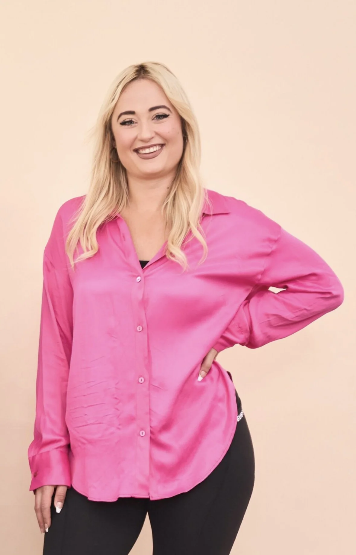 Eine lachende Frau mit blonden Haaren trägt eine pinke Bluse und schwarze Hose.