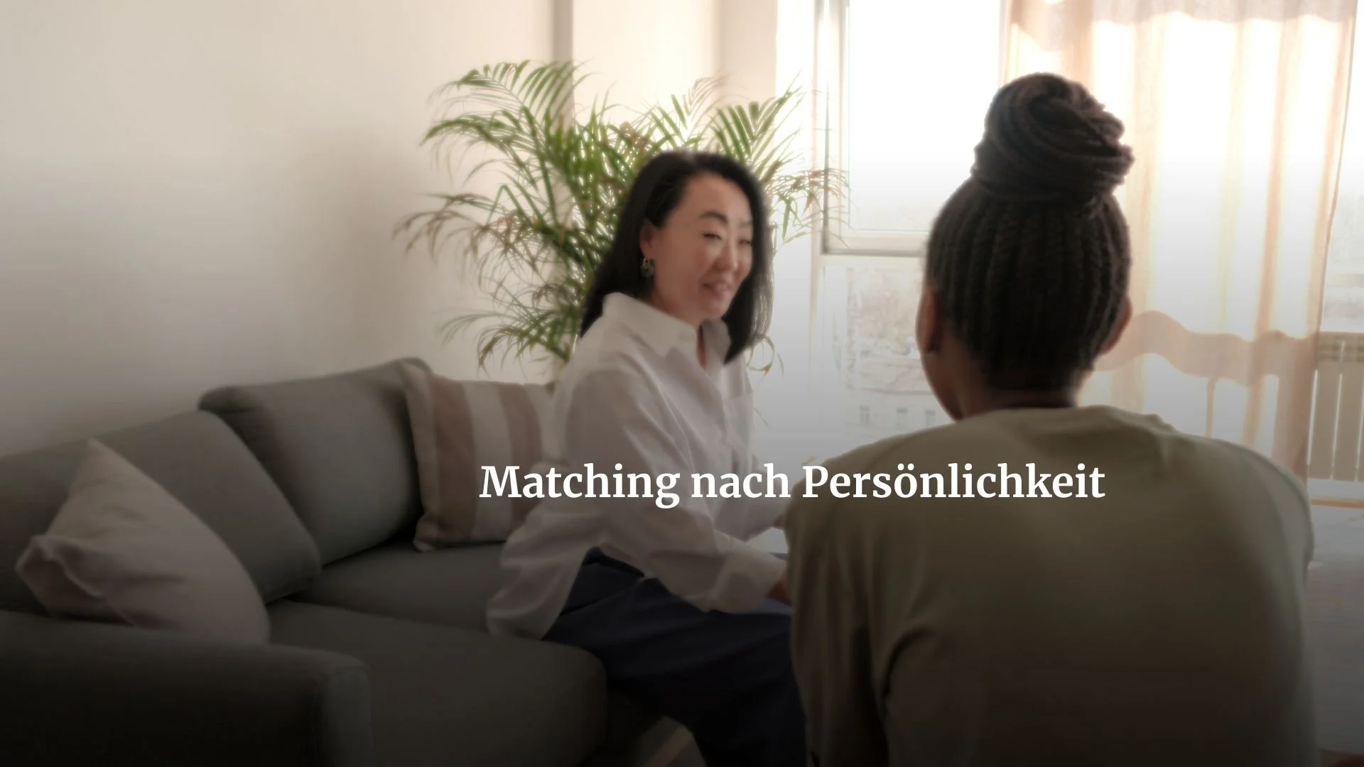 Zwei Frauen sprechen miteinander in einem Wohnzimmer mit Sofa, Kissen und einer großen Pflanze im Hintergrund.