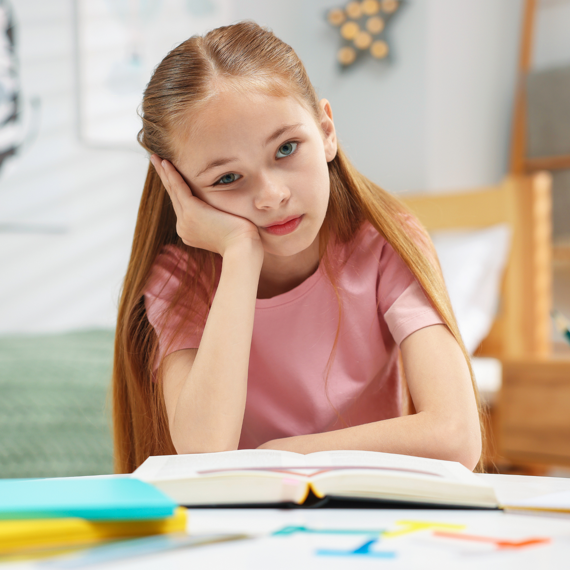 Una ragazza con capelli castani lunghi, occhi azzurri, indossa una maglietta rosa, seduta a un tavolo con libri e quaderni, con un'espressione pensierosa in una stanza ben illuminata.