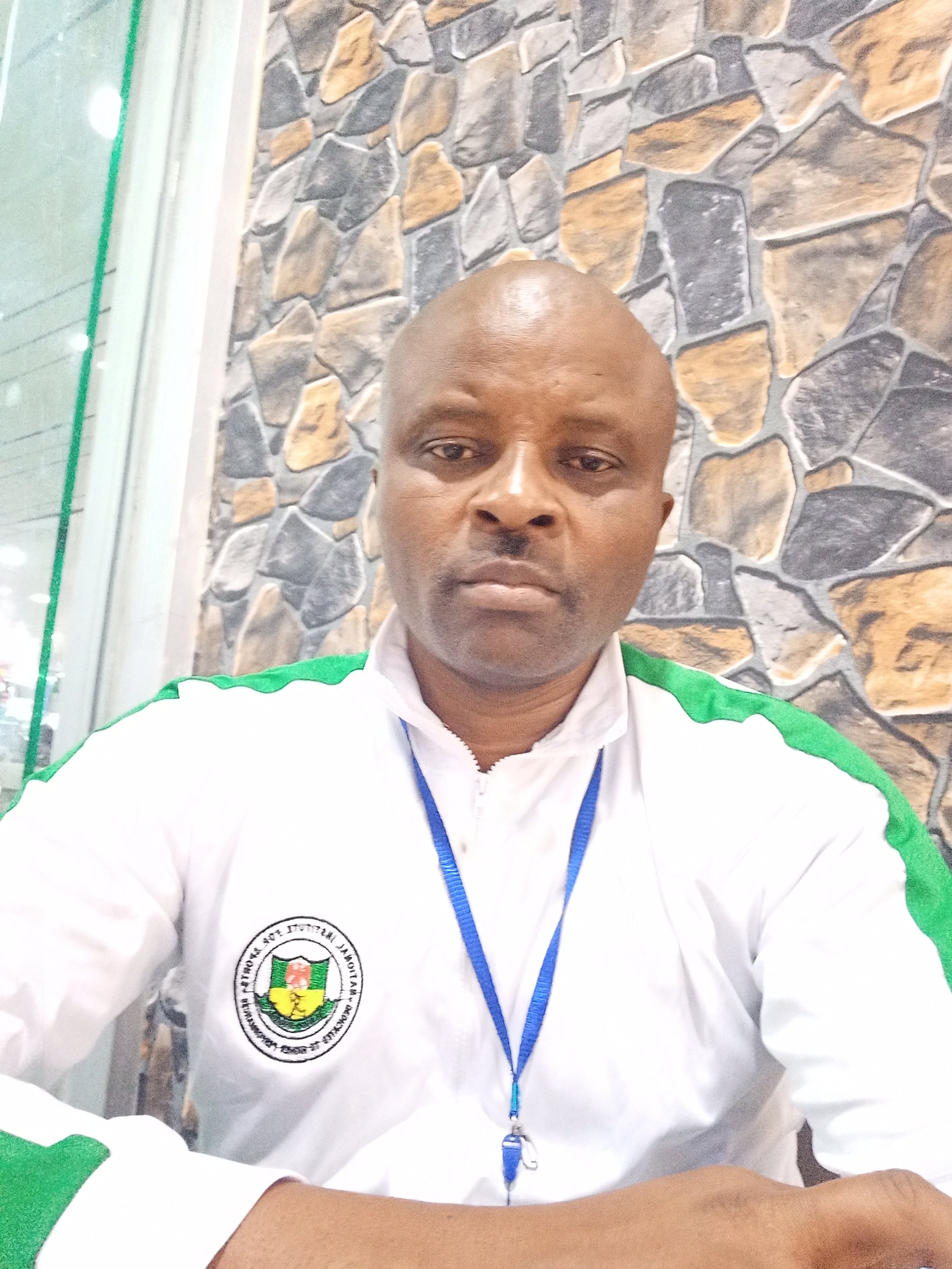 A man taking a selfie in front of a stone wall, wearing a white jacket with green accents and a logo on the chest, and a blue lanyard.